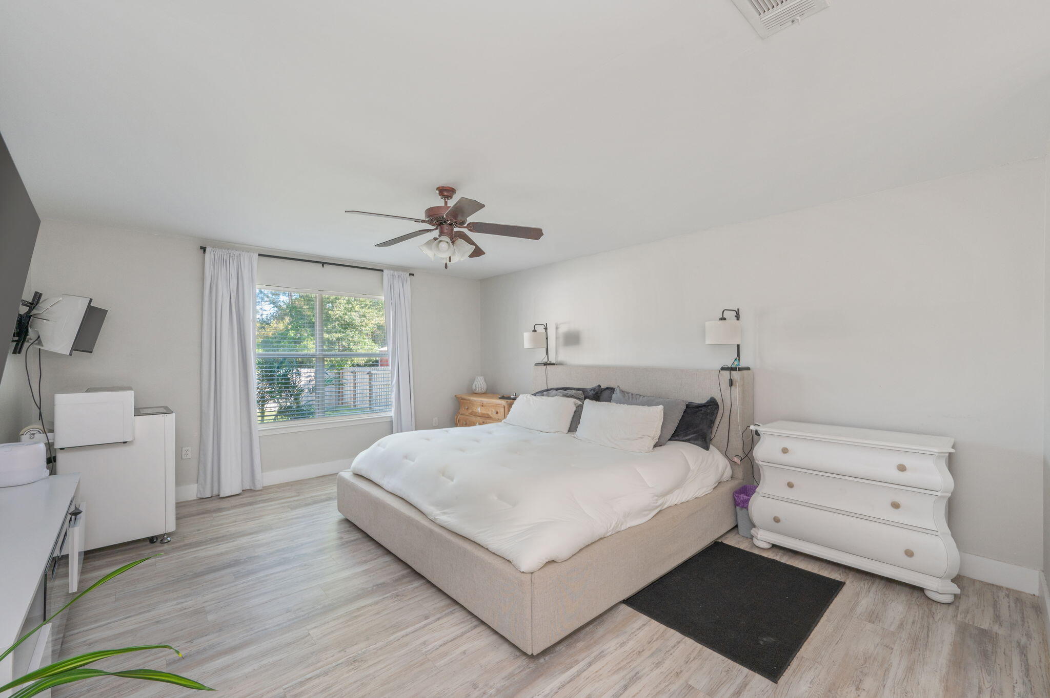 Furnished bedroom with a large white bed, ceiling fan, curtained window, and white dresser.