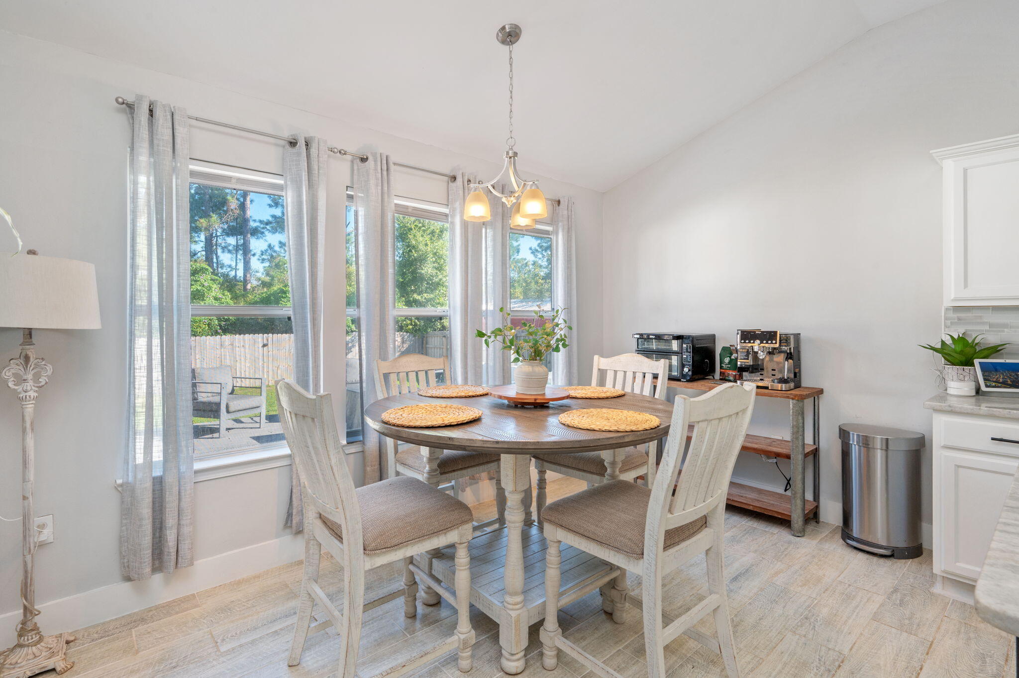 Dining area with a round table, white chairs, and tall windows overlooking a fenced yard.