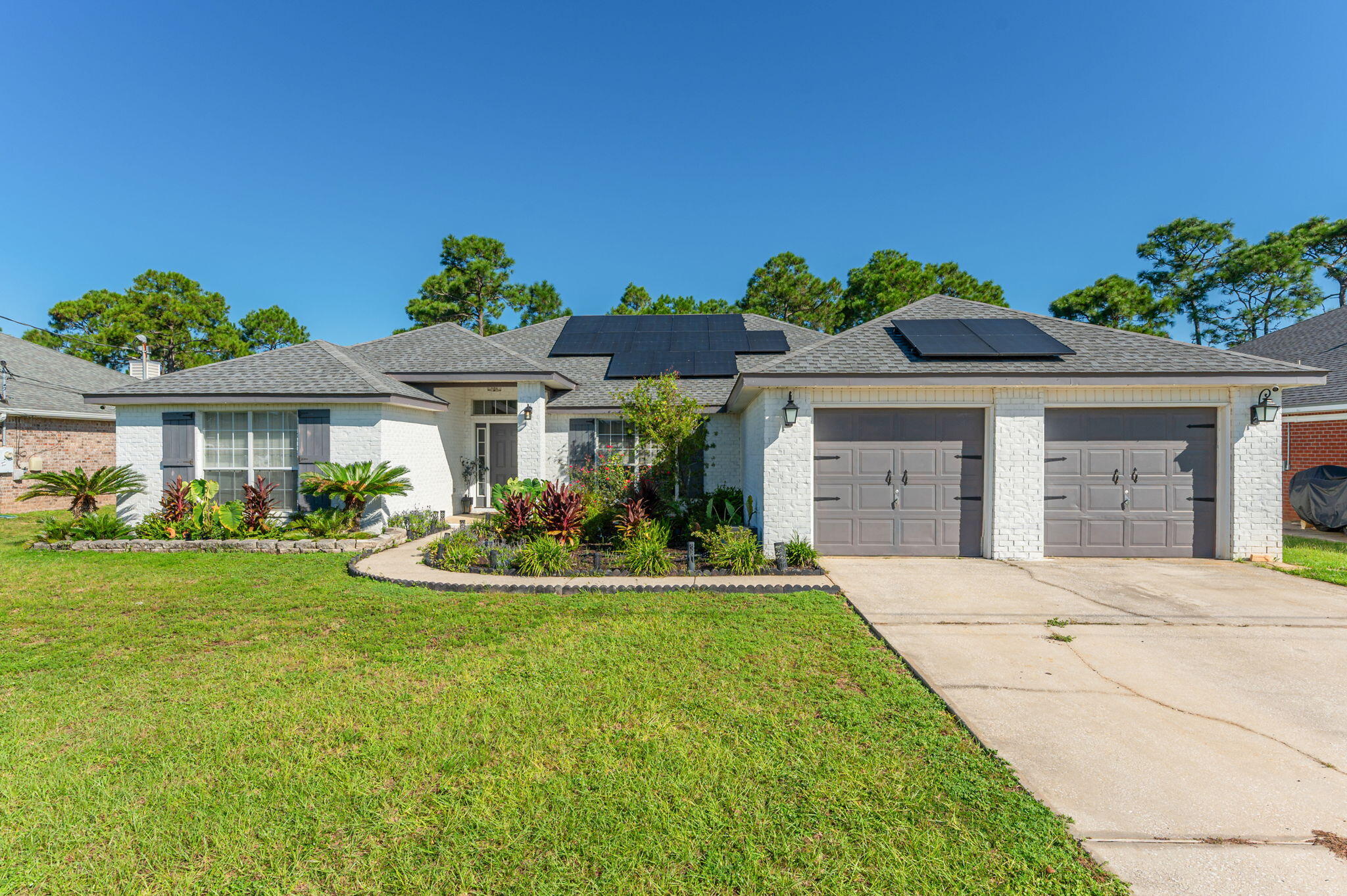 White brick single-story home with rooftop solar panels, a landscaped front garden, and a two-car garage.
