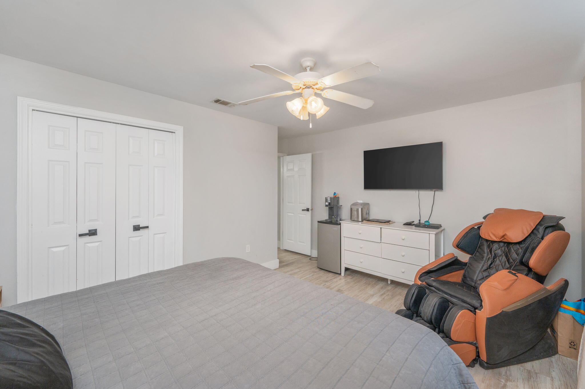 Bedroom with a large bed and a black-and-orange massage chair beside a dresser and wall-mounted TV.