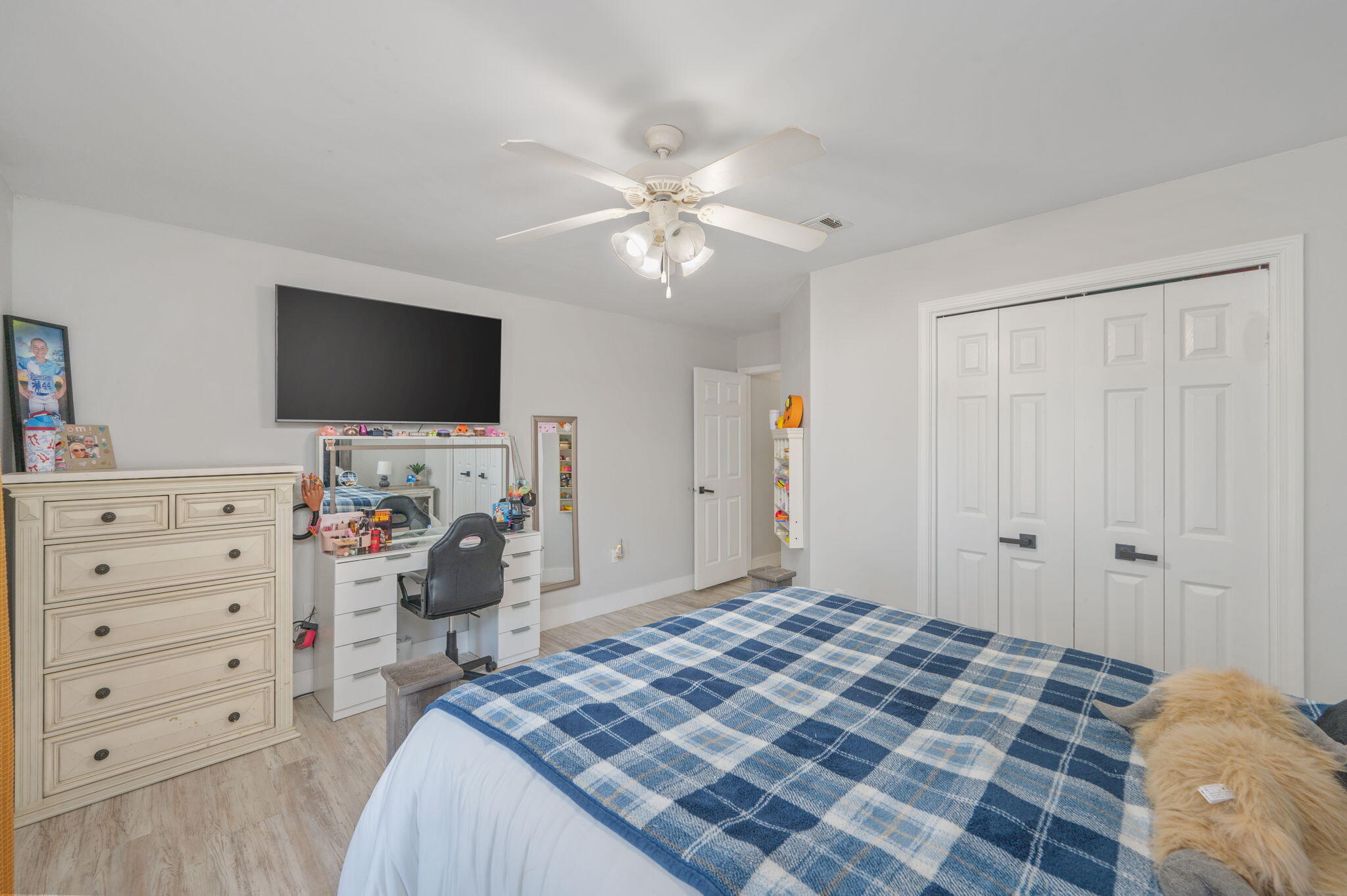 Bedroom with a blue plaid bedspread, ceiling fan, dresser, desk, and white closet doors.