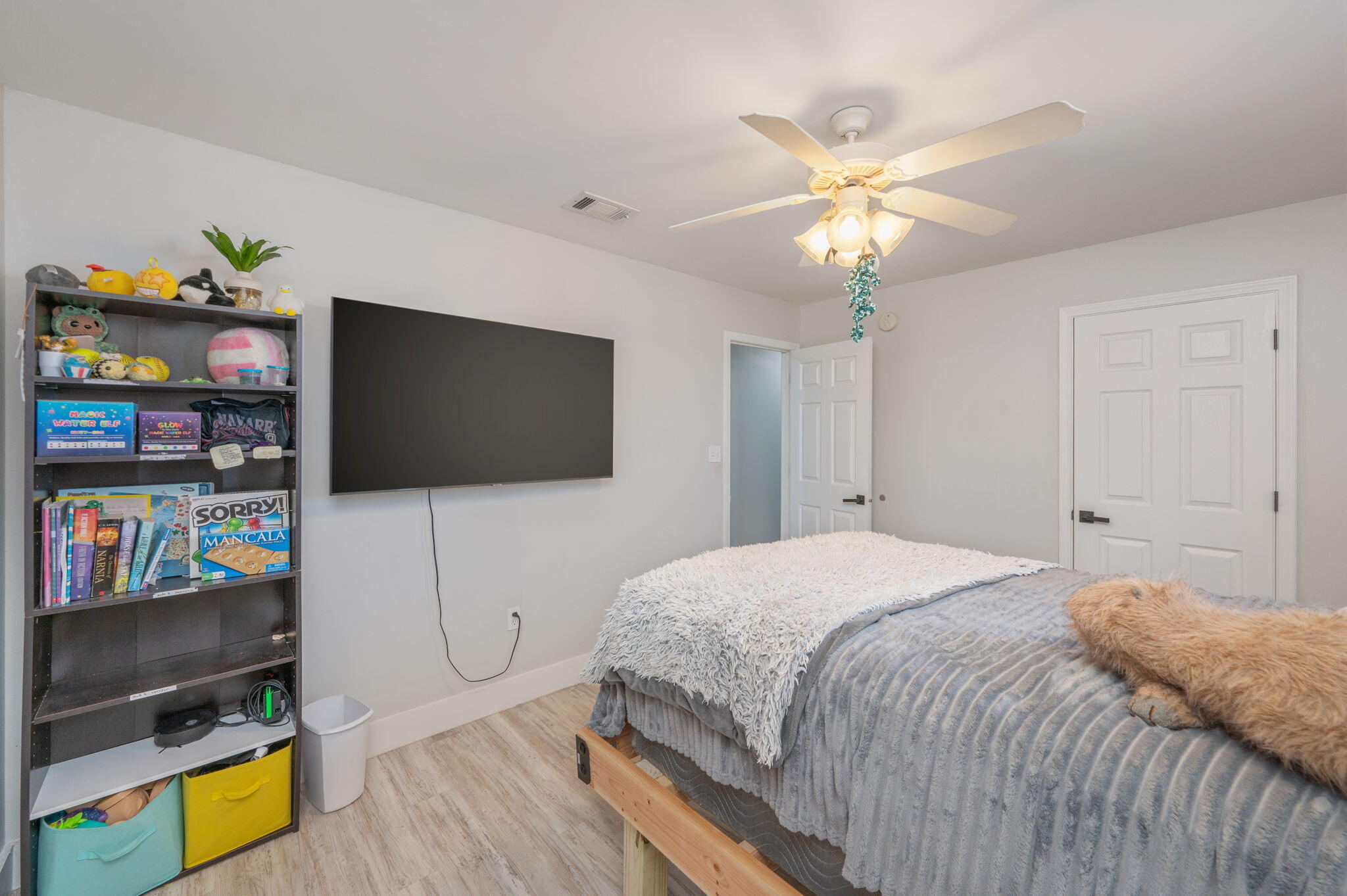 Bedroom with gray bedding, a fluffy white throw, wall-mounted TV, and toy-filled bookshelf.