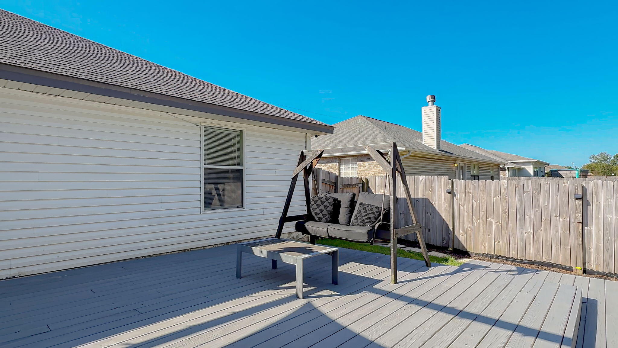 Backyard deck with a wooden swing bench and outdoor table beside a white-sided house.