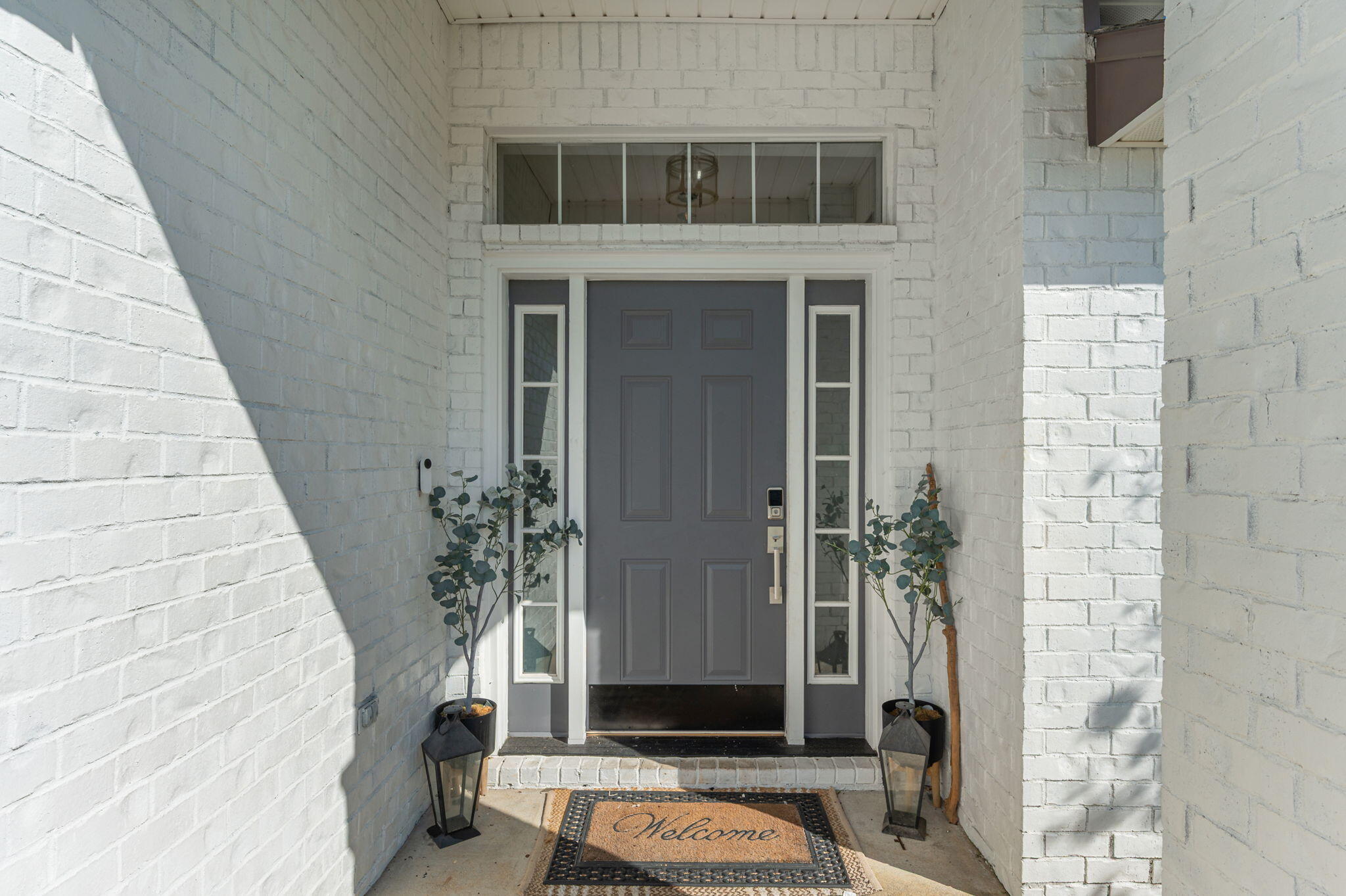 Covered front entry with a gray paneled door, white brick walls, potted plants, and a welcome mat.