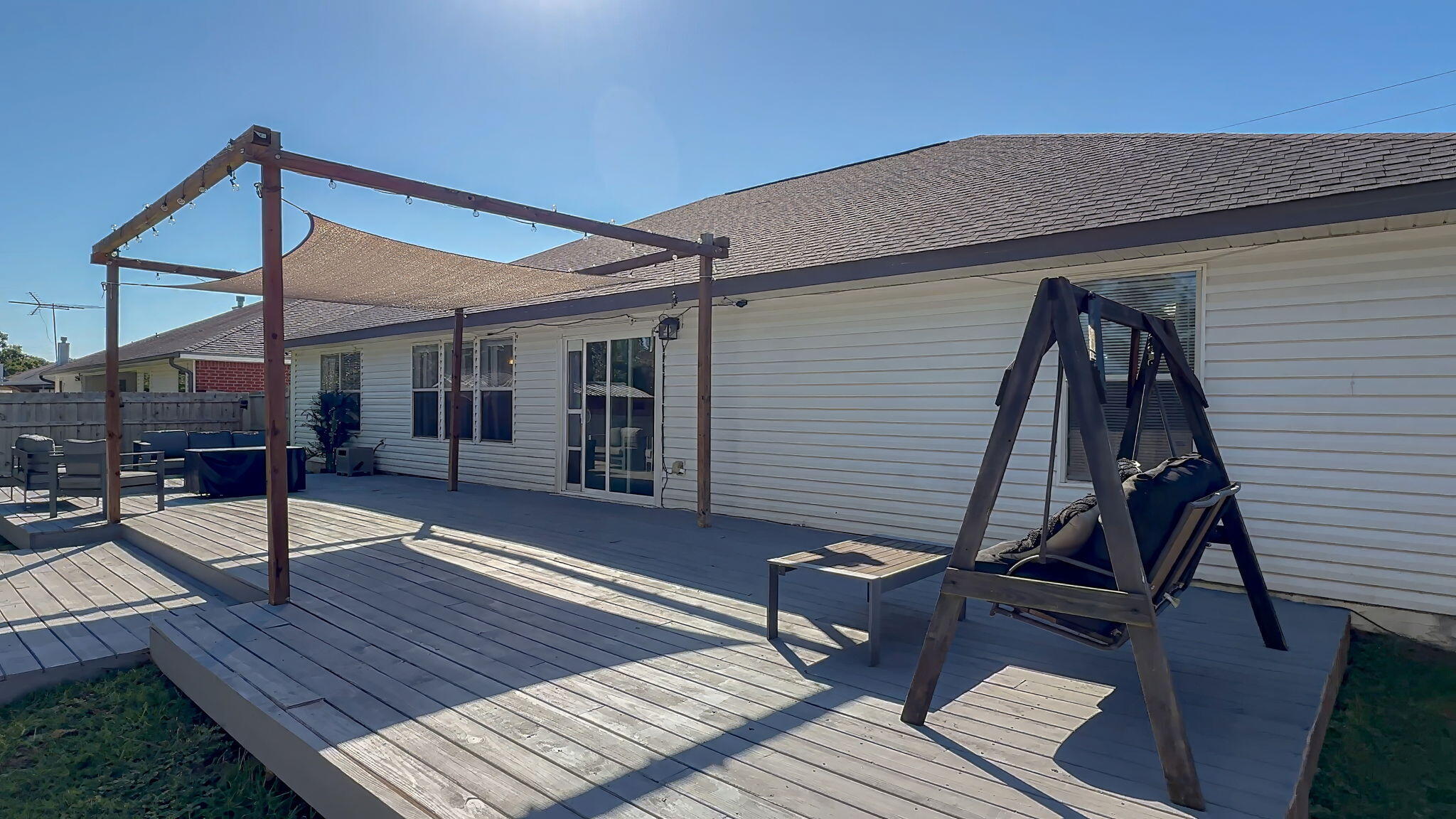Raised wooden deck with pergola, shade sail, patio seating, and swing beside a white-sided home.