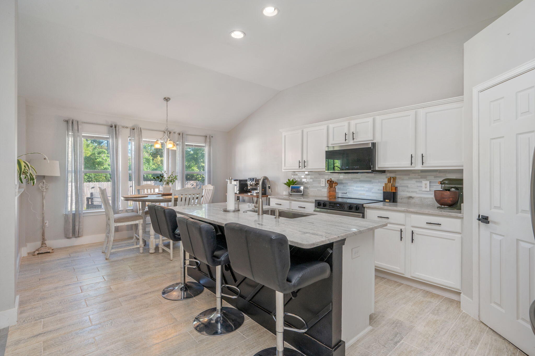 Kitchen with a large island and dark bar stools beside a dining nook and curtained windows.