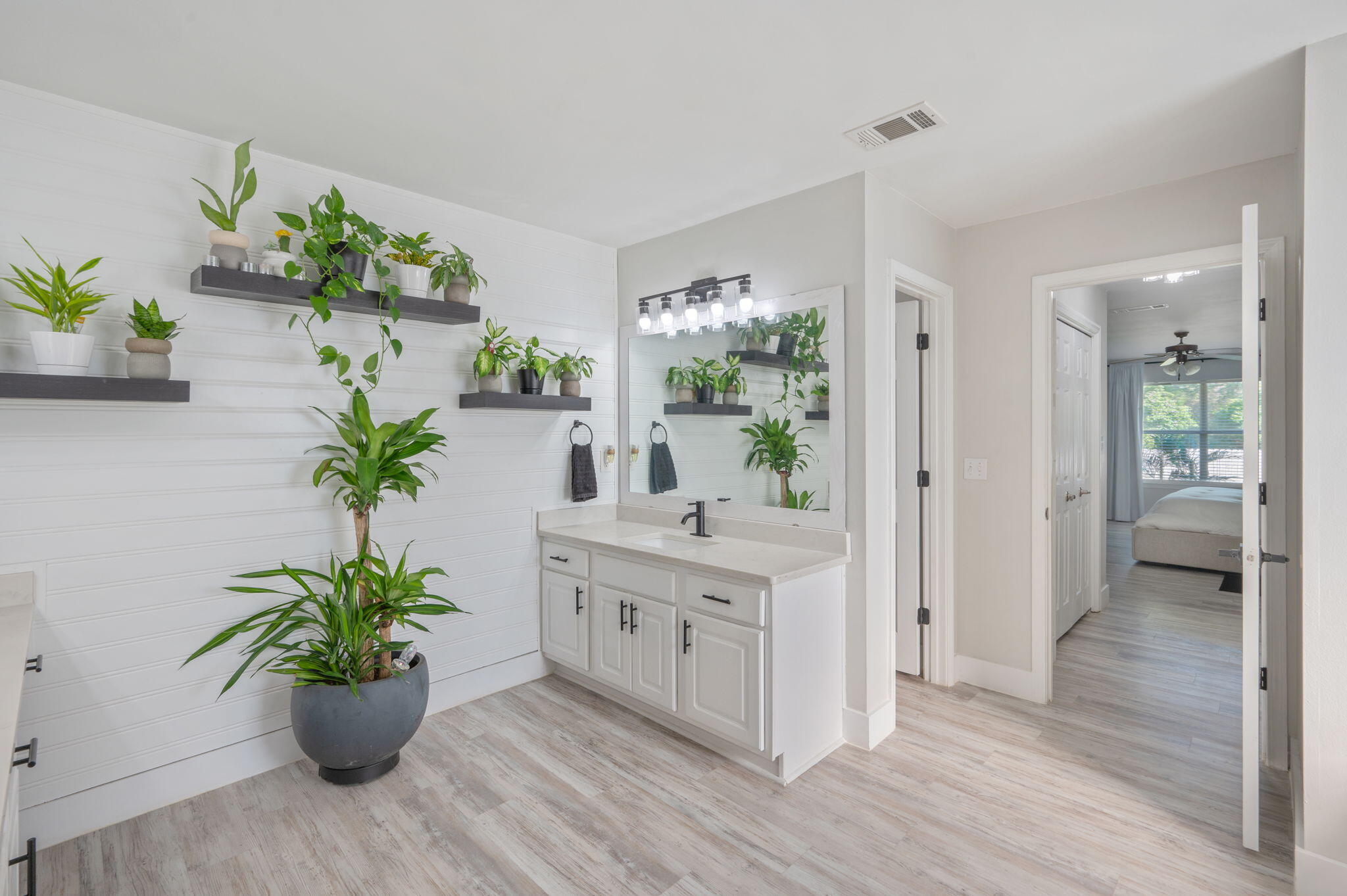 Bright bathroom vanity with a large mirror, plant-lined shelves, and an open doorway to a bedroom.