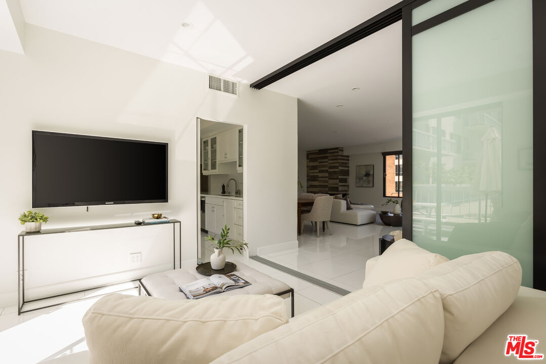Living room with white sofas, a wall-mounted television, and a glass partition opening to the kitchen.