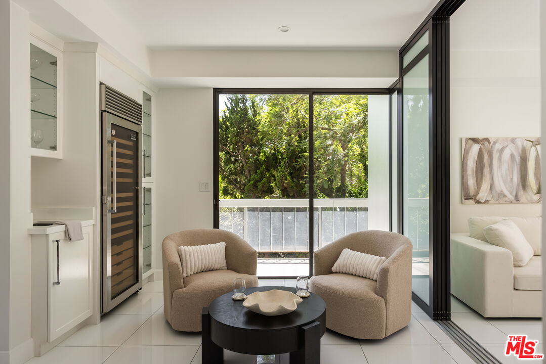 Living area with beige armchairs, a black coffee table, and sliding doors to a leafy balcony.