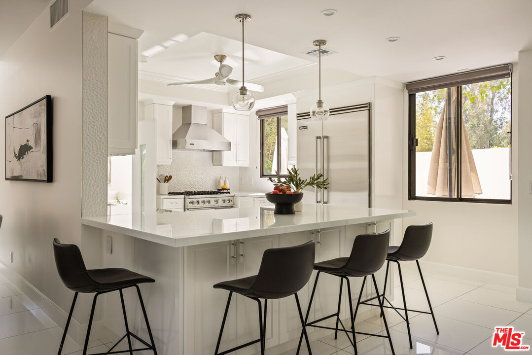 Modern white kitchen with a large island, four black stools, stainless steel appliances, and leafy views.