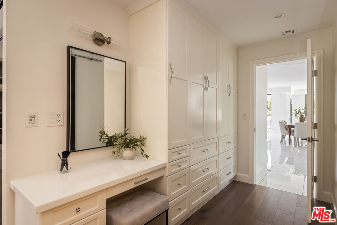 Dressing area with white built-in cabinets, a vanity, and an upholstered bench beside an open doorway.