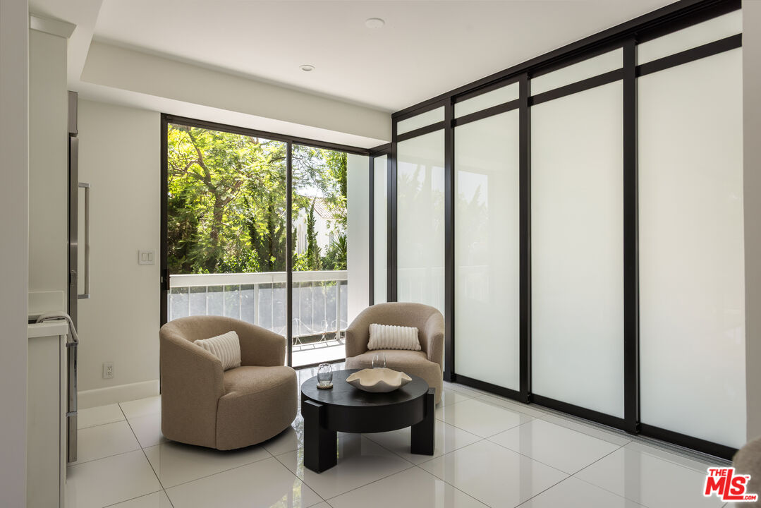 Living area with beige armchairs, black coffee table, frosted-glass panels, and a leafy view through the window.
