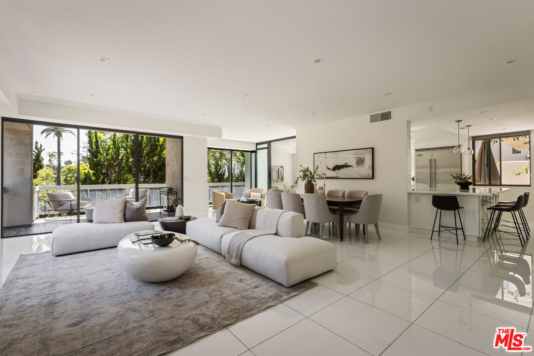 Open-plan living and dining room with white sofas, glossy tile floors, and a balcony overlooking trees.