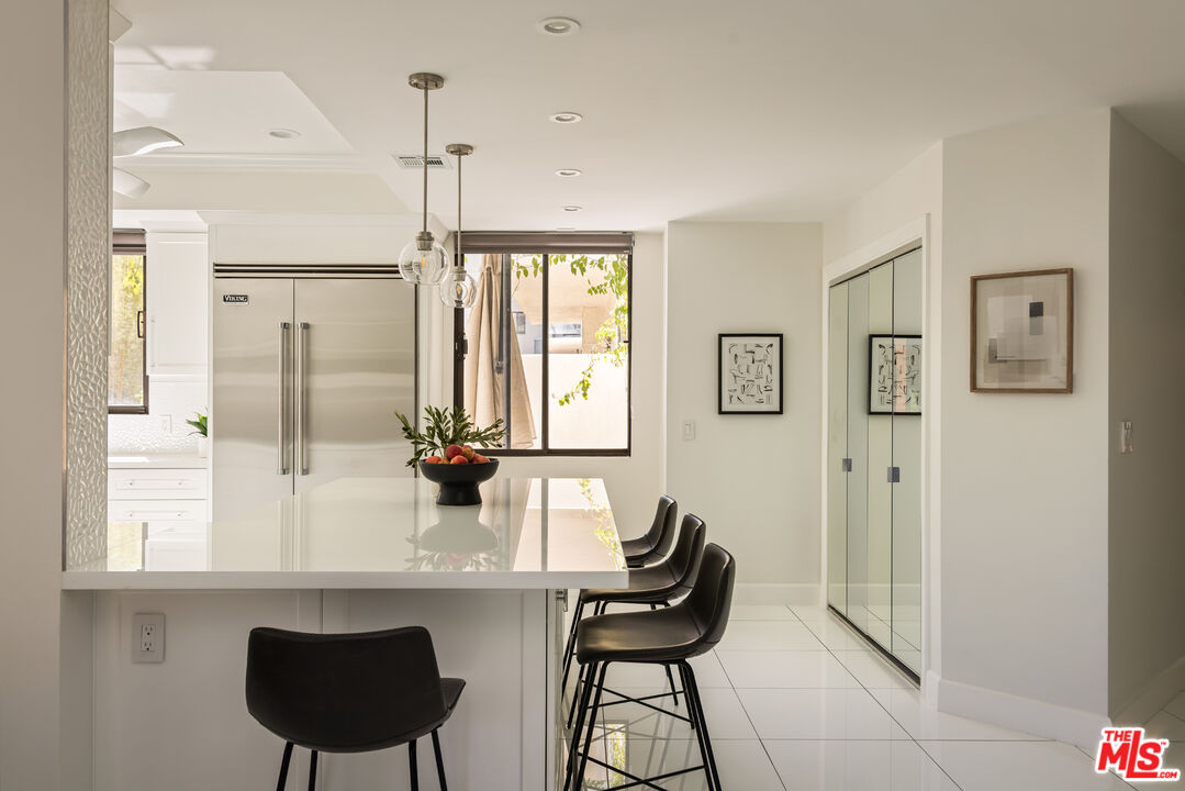 Modern kitchen with white island, black bar stools, and stainless-steel refrigerator beside large windows.