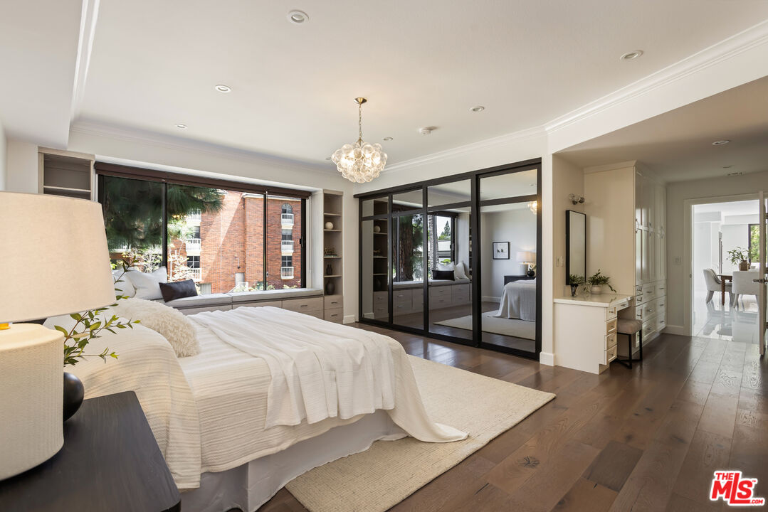 Furnished bedroom with a white bed, mirrored closet doors, and a large window overlooking a brick building.