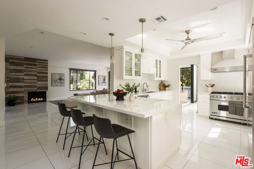 Open-plan kitchen with a large white island, dark bar stools, stainless steel range, and adjoining fireplace.