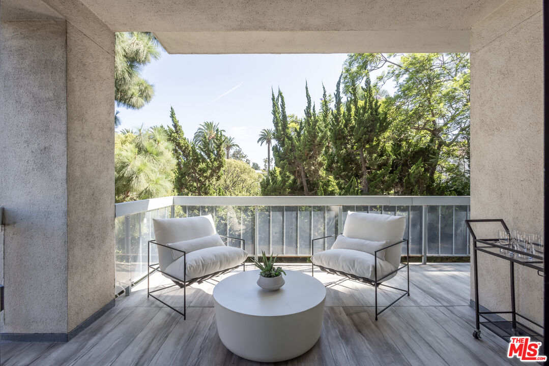 Apartment balcony with white lounge chairs and a round table overlooking leafy trees.
