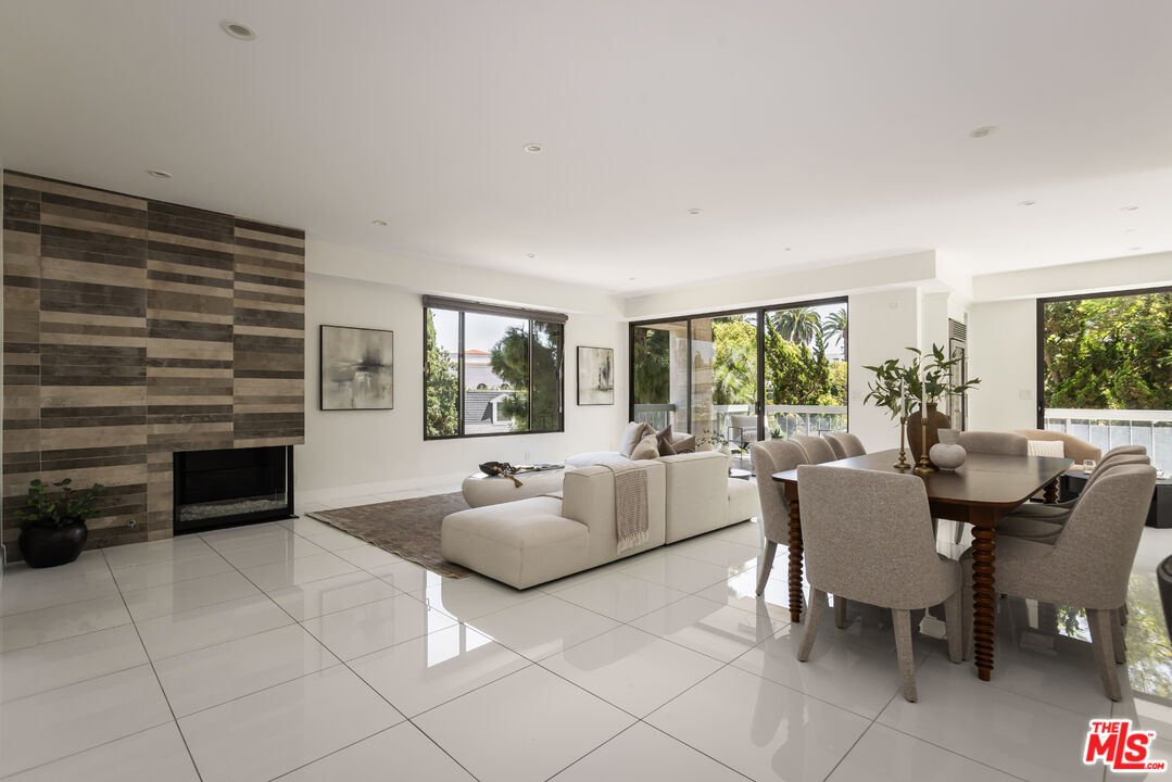 Open-plan living and dining room with a striped stone fireplace wall and large windows overlooking greenery.