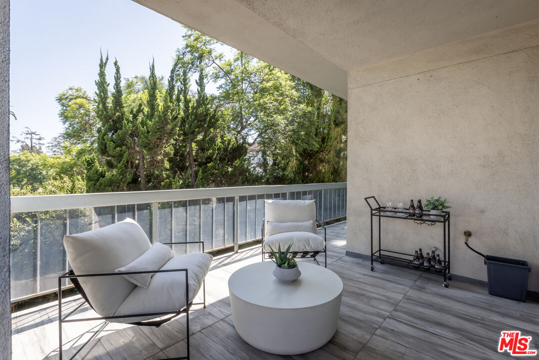 Apartment balcony with white lounge chairs and a round coffee table overlooking trees.