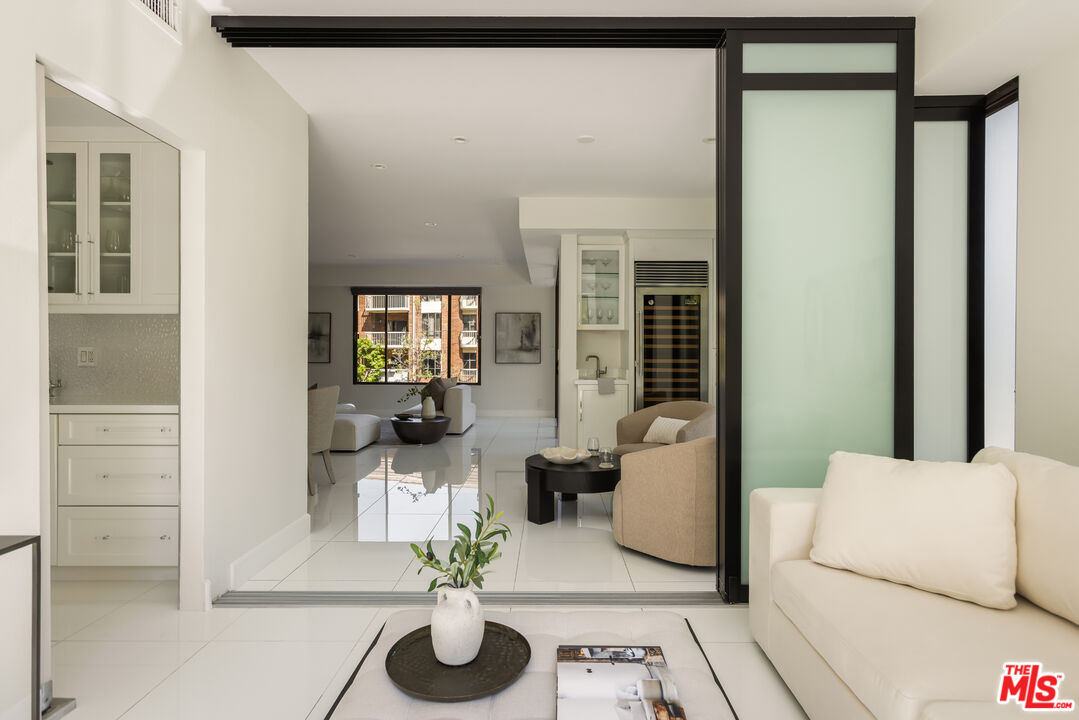 Staged condo living room with cream sofas, glossy white tile floors, and a black-framed frosted glass partition.