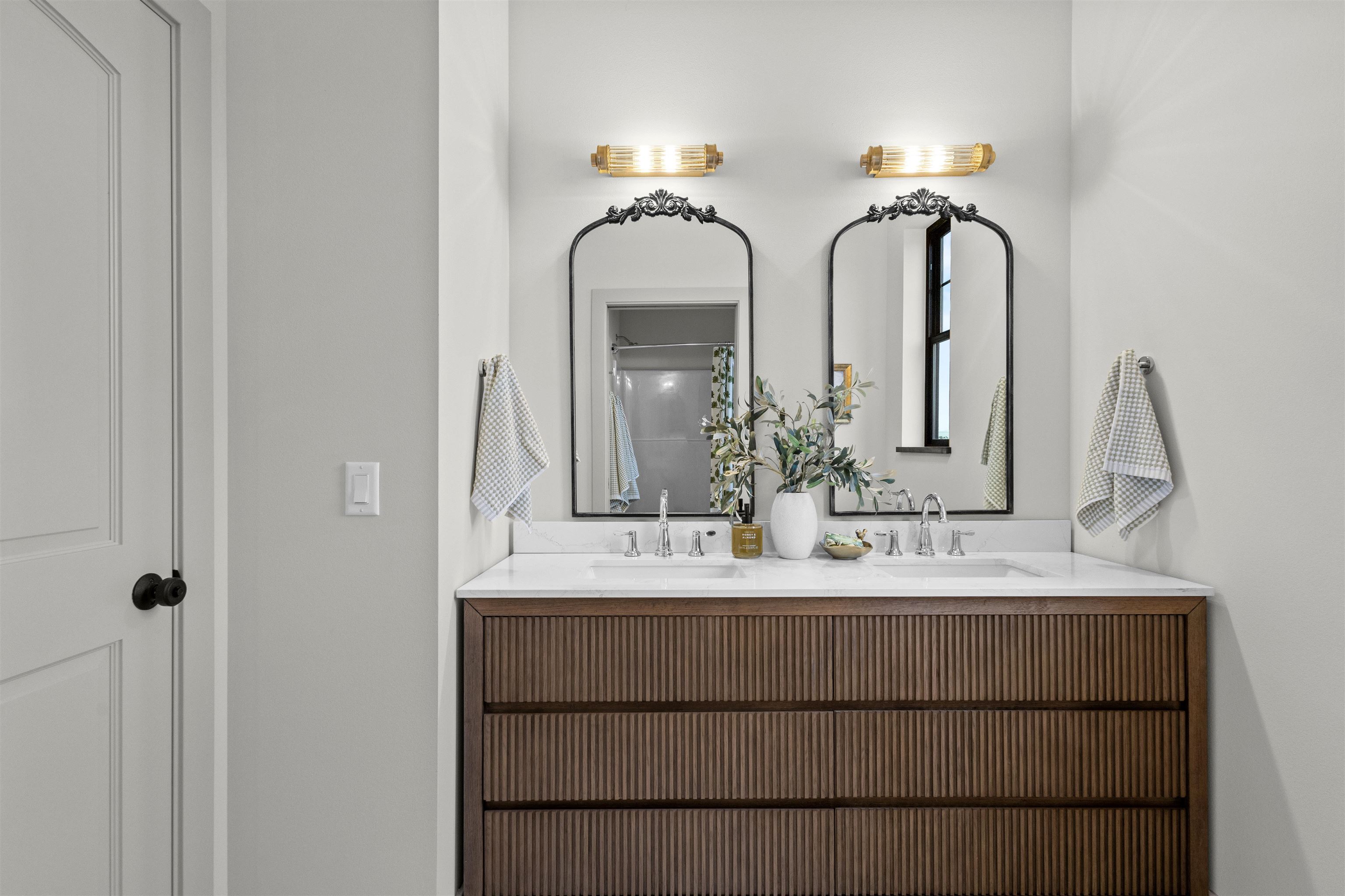 Bathroom with a double-sink ribbed wood vanity, ornate mirrors, and brass wall lights.