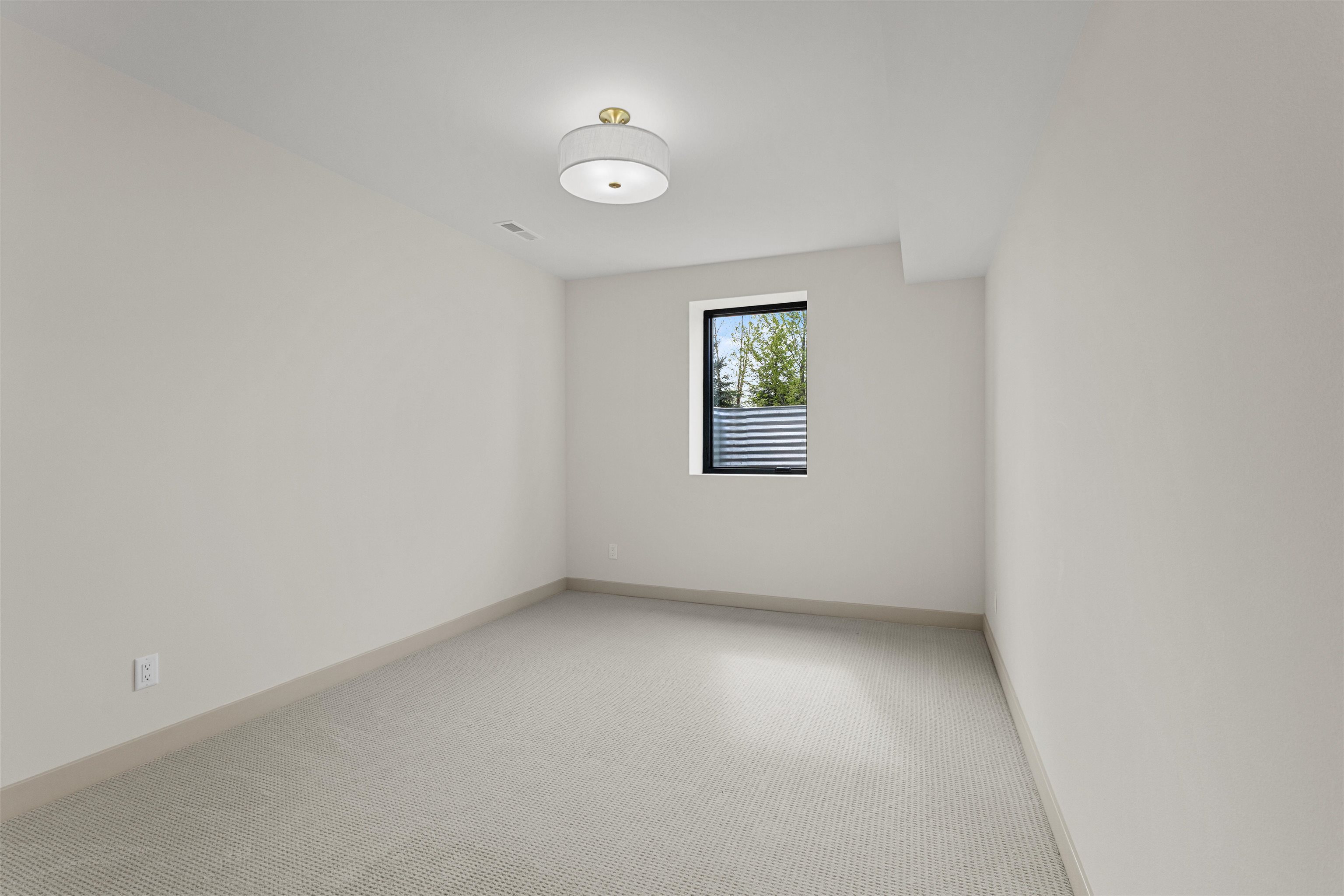Empty carpeted room with a small window overlooking trees and a metal fence.