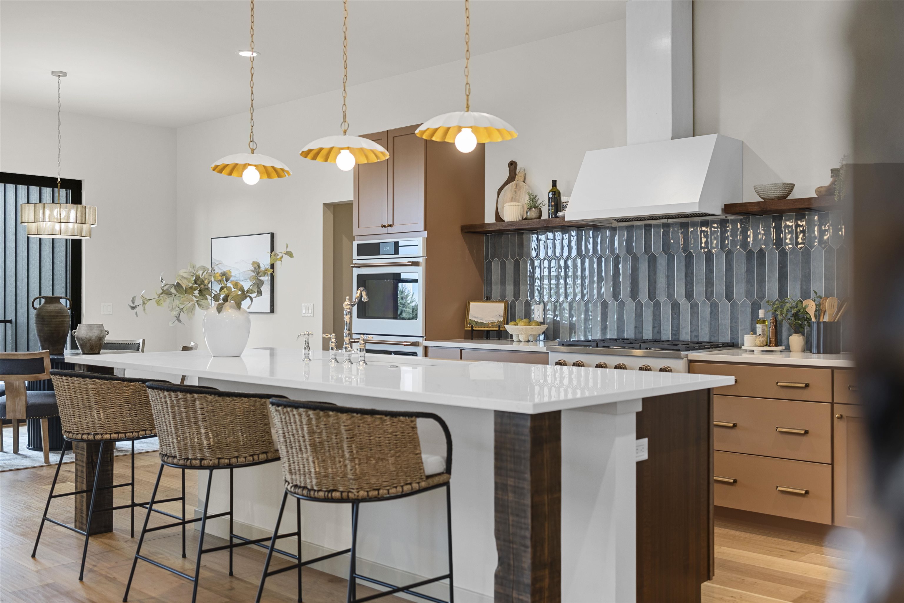 Contemporary kitchen with a white waterfall island, woven stools, dark glossy tile backsplash, and warm brown cabinets.