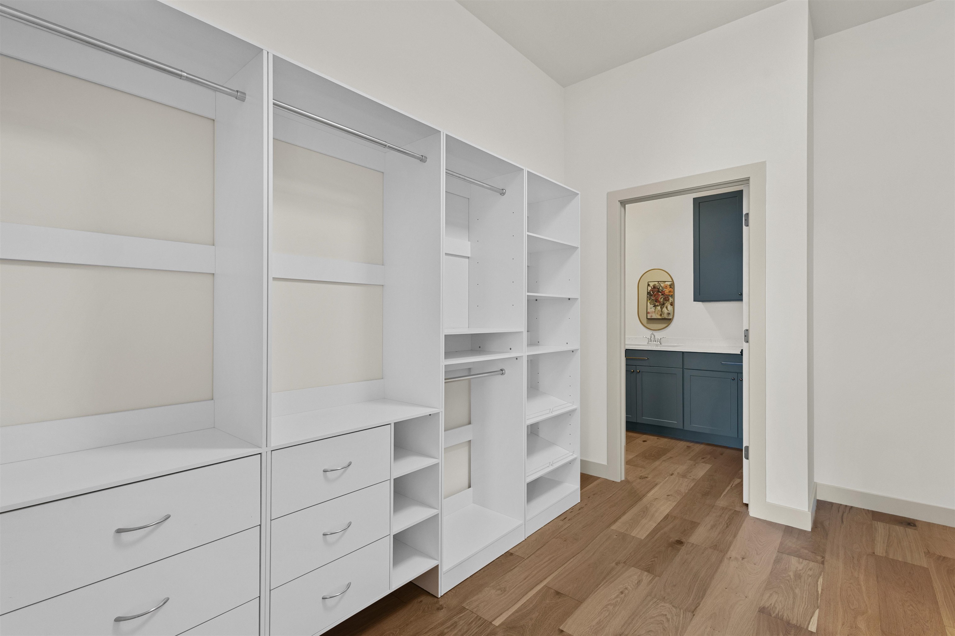 Walk-in closet with white built-in storage and a doorway to a bathroom with a dark blue vanity.
