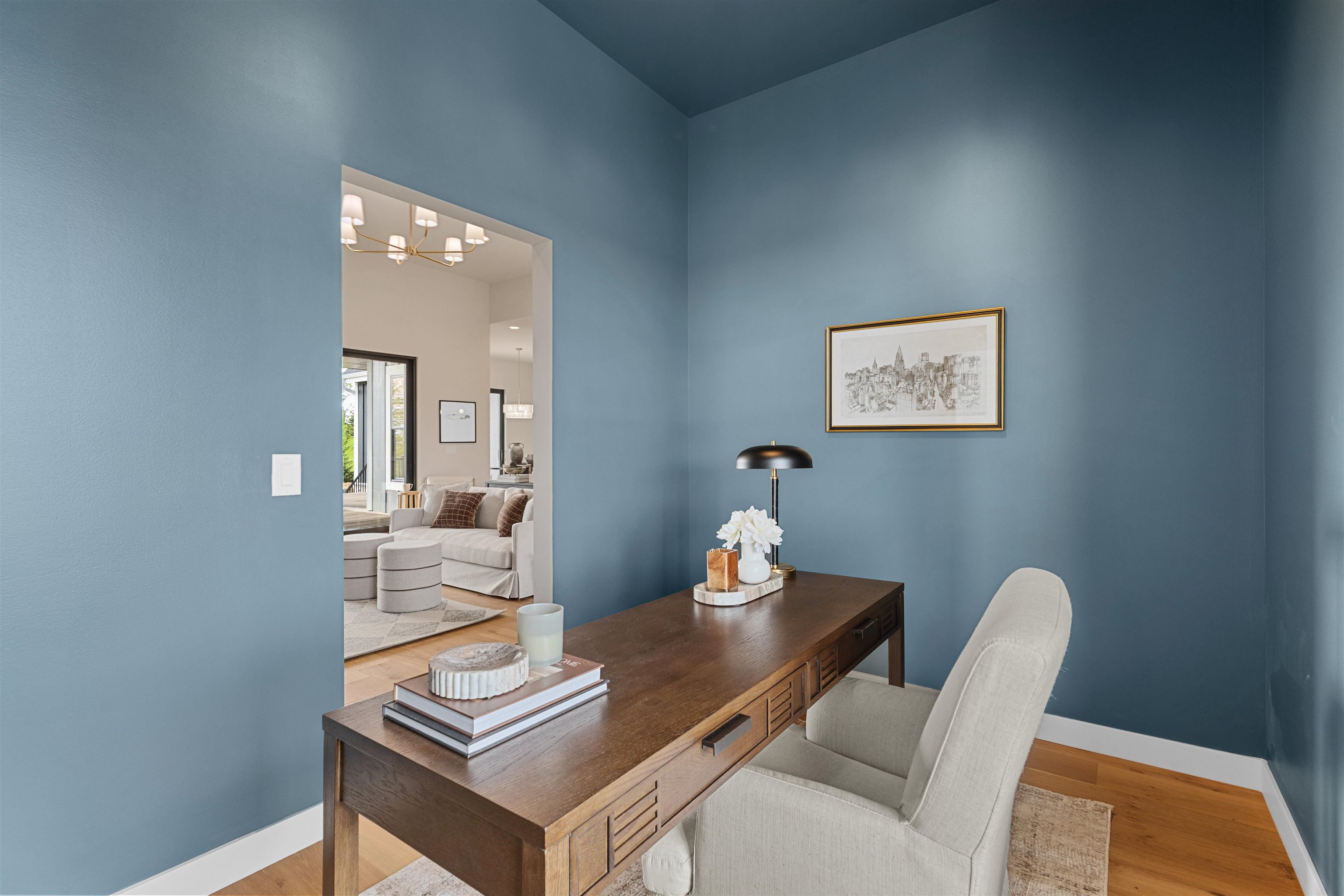 Home office with blue walls, a dark wood desk, and a white chair opening onto a bright living room.
