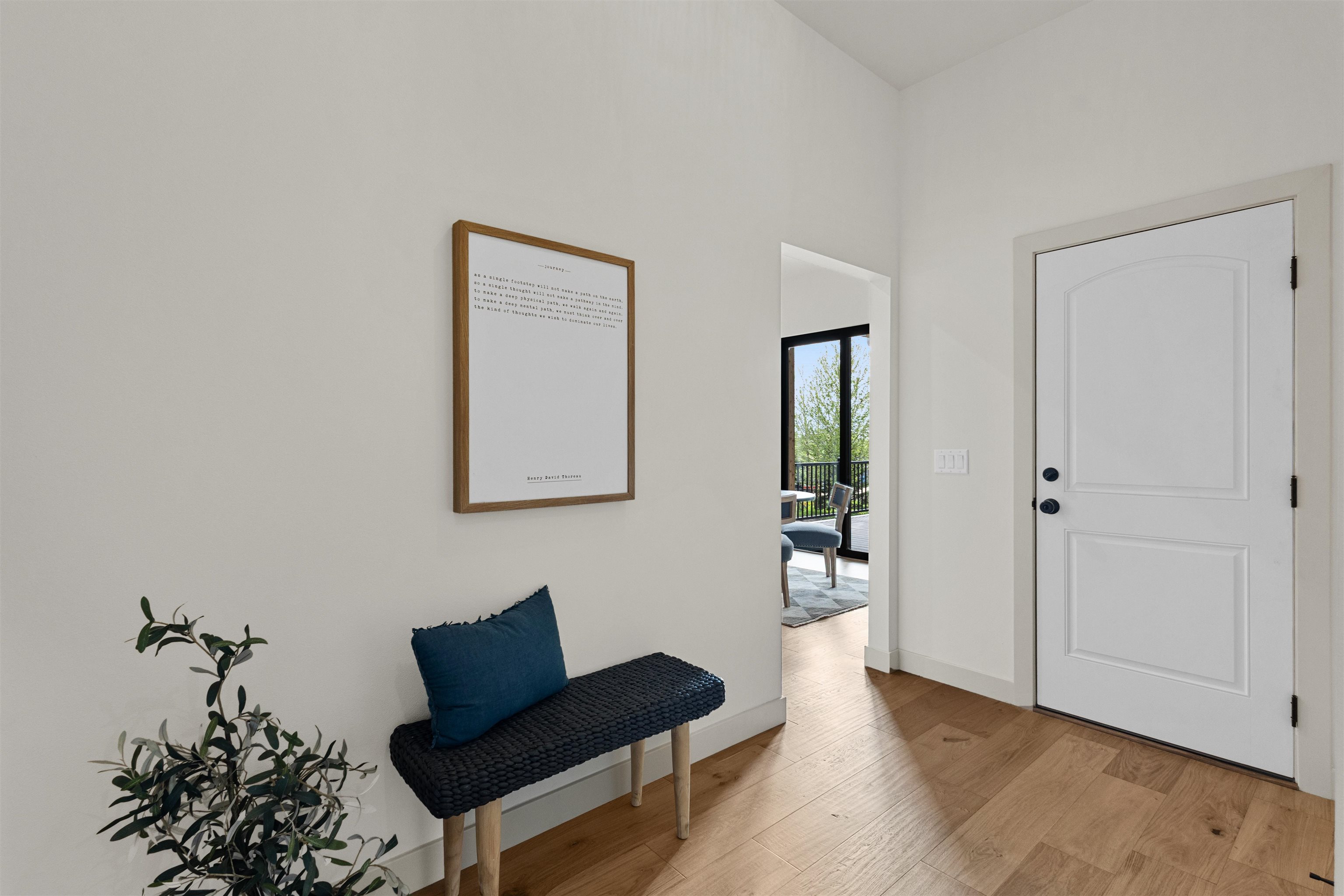 Entryway with a white front door, woven bench, blue cushion, potted plant, and framed quote.