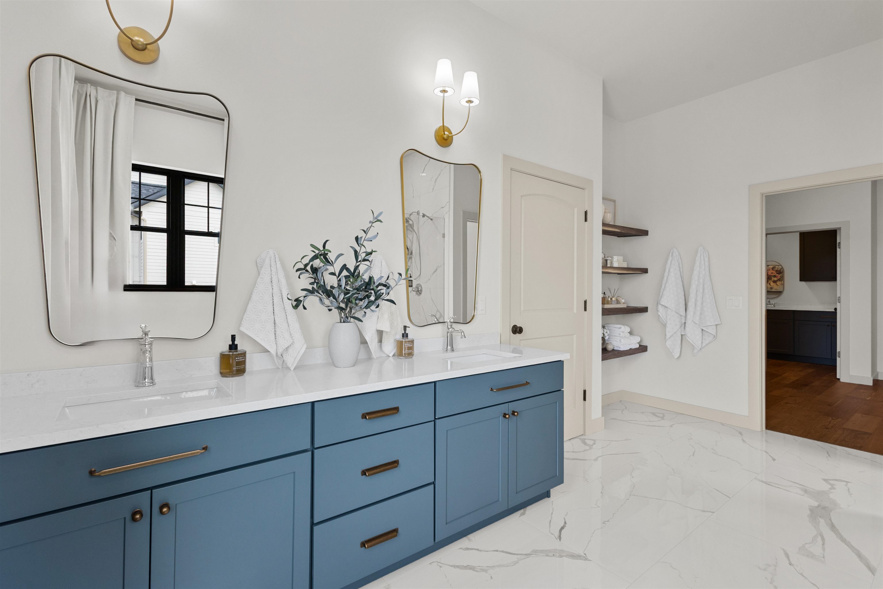 Bright bathroom with a blue double vanity, brass fixtures, gold-framed mirrors, and marble-look floors.