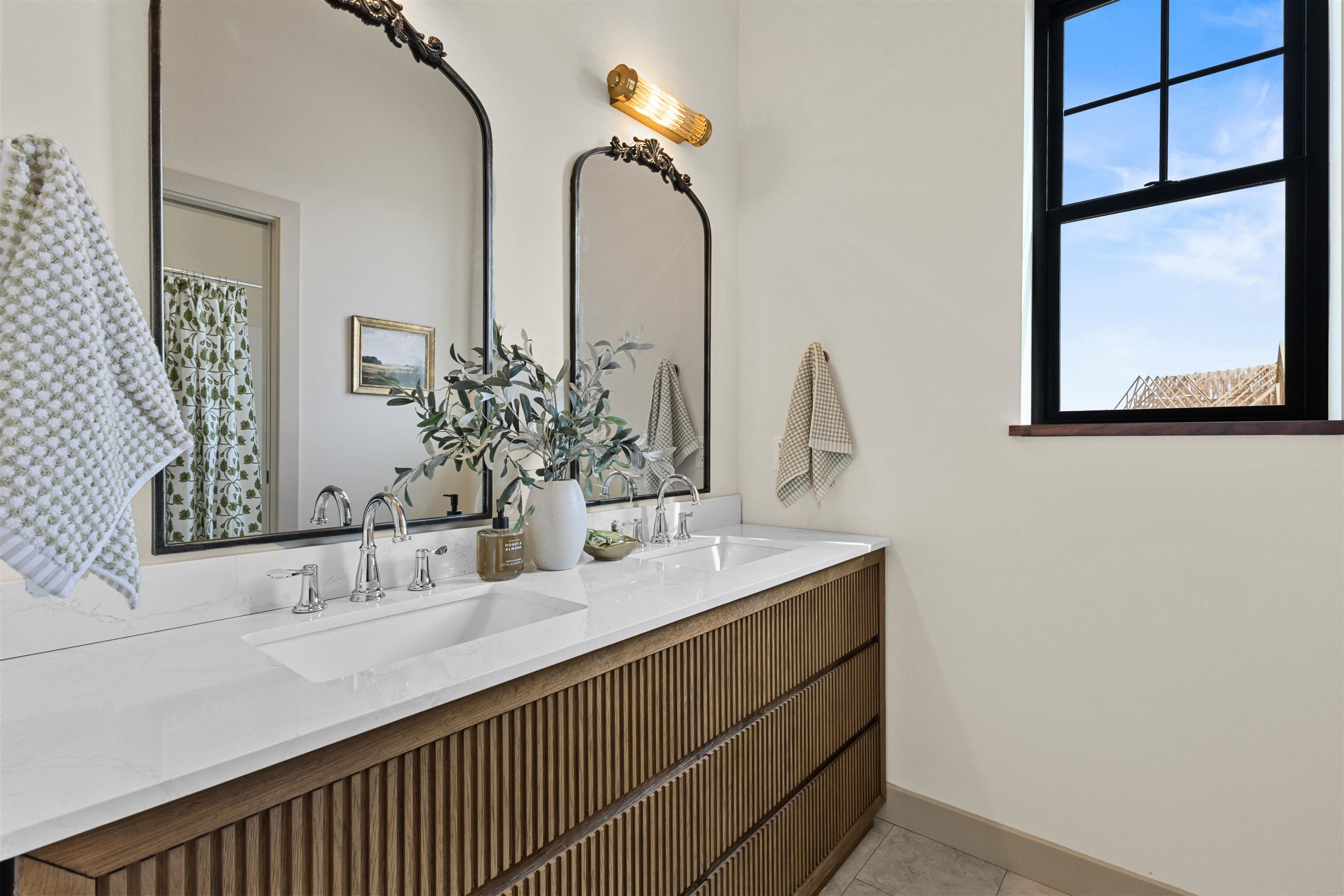 Bathroom with a fluted wood double vanity, white stone counter, ornate mirrors, and a large black-framed window.