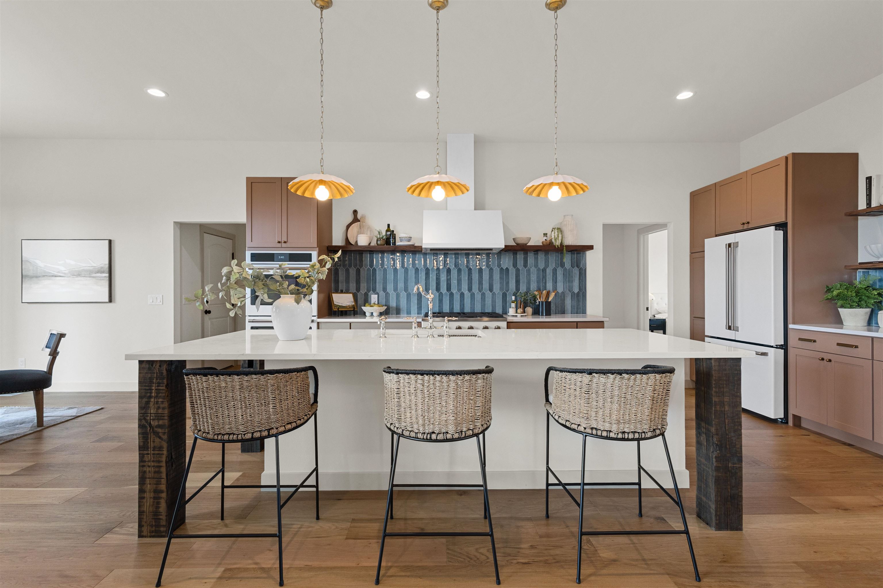 Modern kitchen with a large white island, woven bar stools, blue tile backsplash, and warm brown cabinetry.