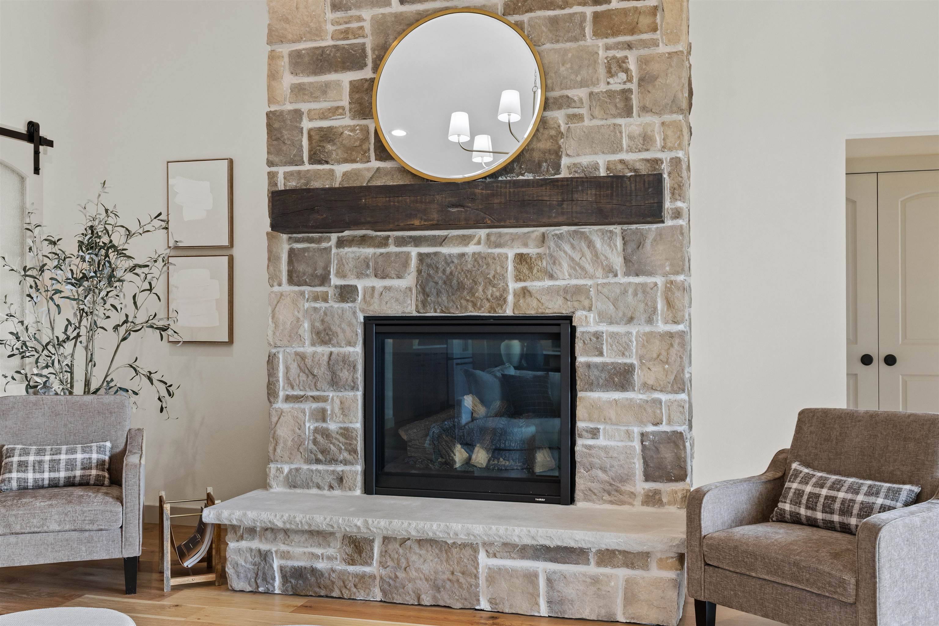 Stone fireplace with a dark wood mantel and round mirror between two upholstered armchairs.