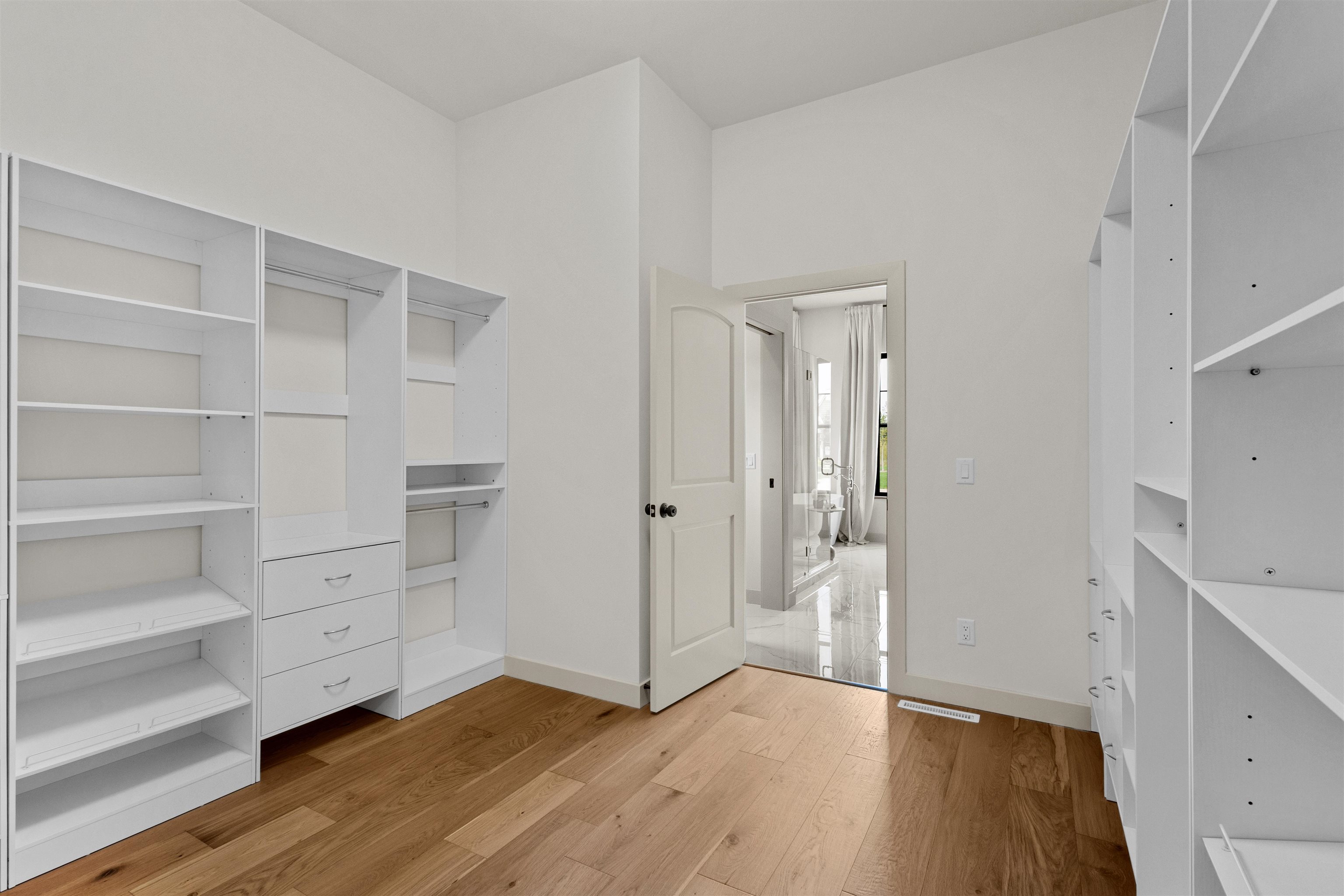 Walk-in closet with white built-in shelving and drawers, wood floors, and an open doorway to a bathroom.