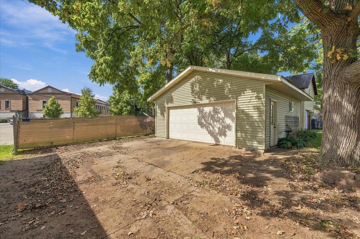 Detached beige garage with a white double door beneath mature trees, beside a concrete driveway.