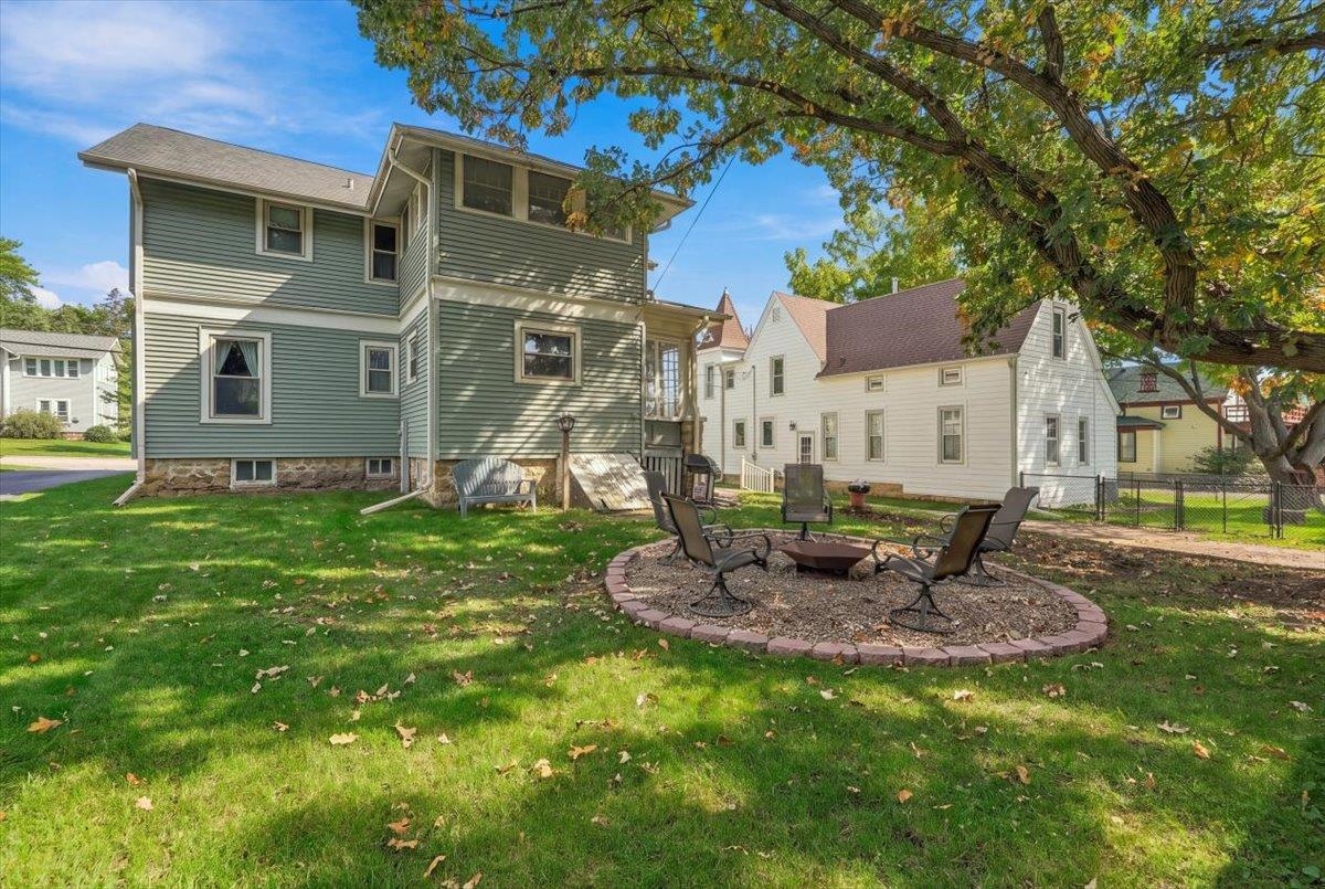 Two-story green home and backyard with a circular fire-pit seating area beneath a large tree.