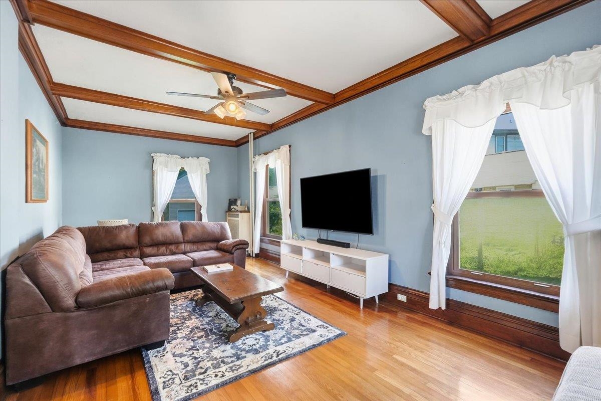 Living room with a brown leather sectional, exposed wood ceiling beams, and tall curtained windows.