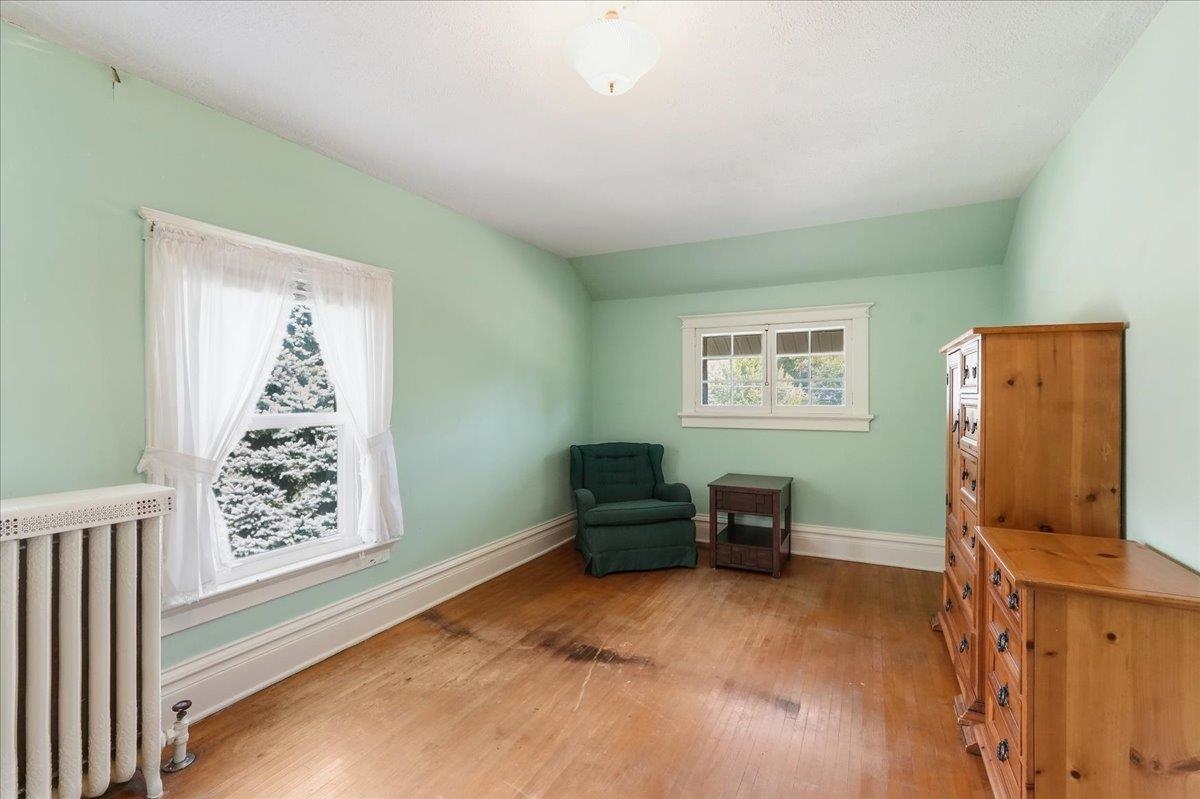 Sparsely furnished mint-green room with hardwood floors, two windows, an armchair, and wooden dressers.