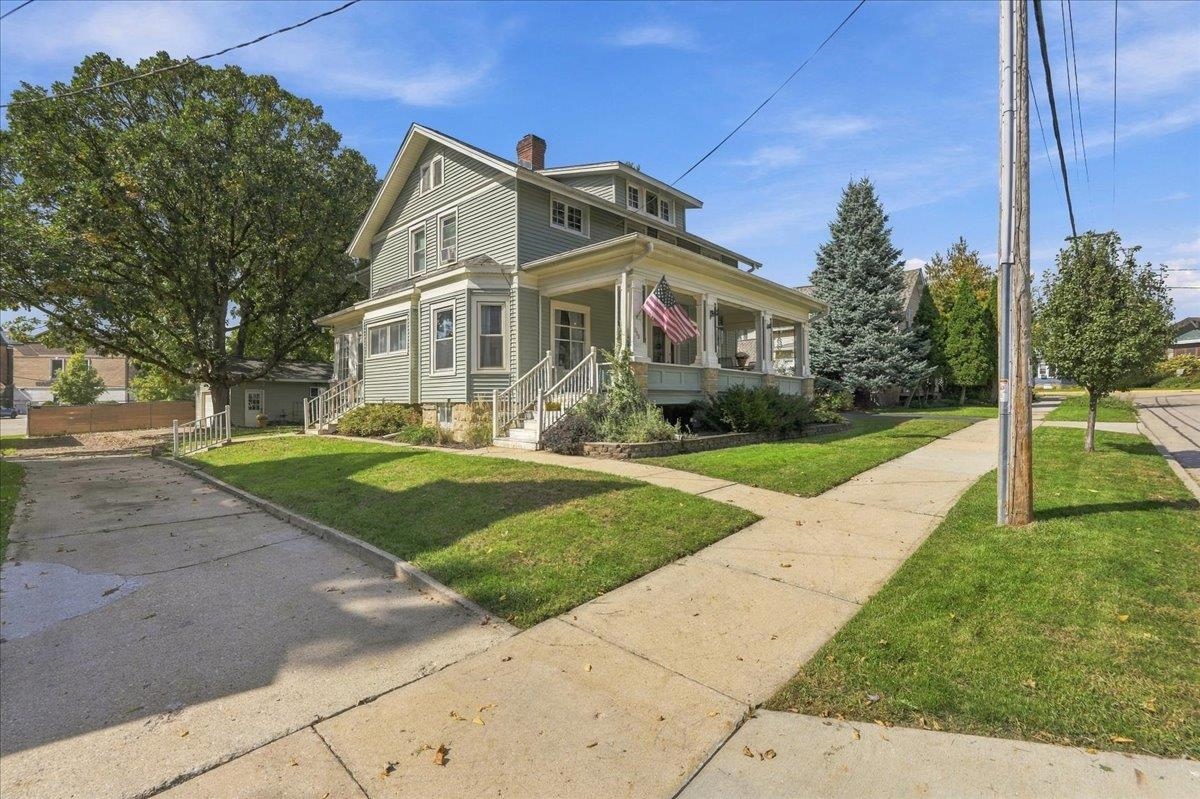 Two-story pale green house with a broad covered porch and American flag on a sunny corner lot.
