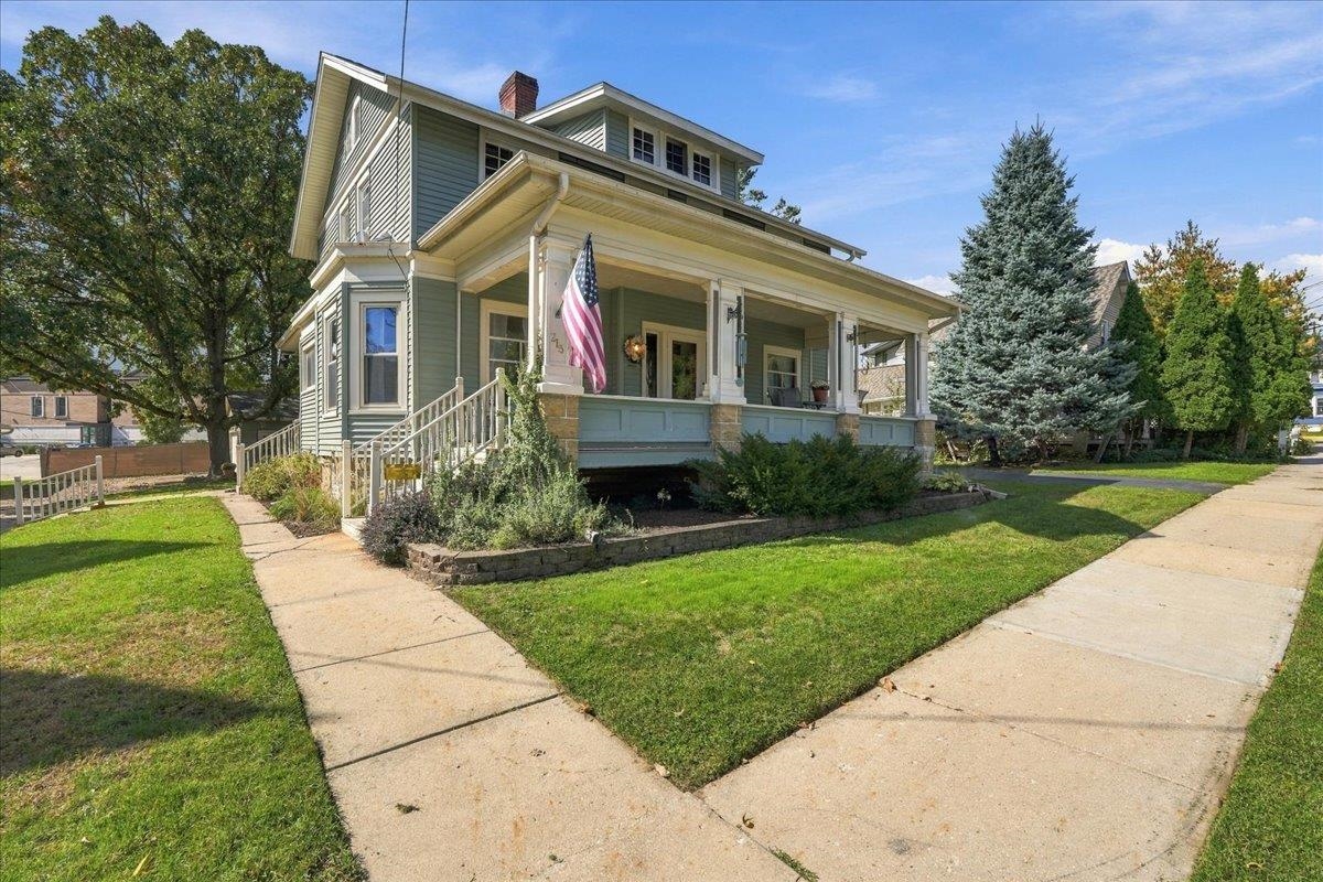 Sage-green two-story home with a broad covered porch, white trim, and an American flag.