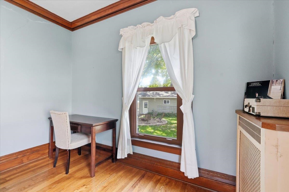 Furnished room with a dark wood desk beside a tall curtained window overlooking a green yard.