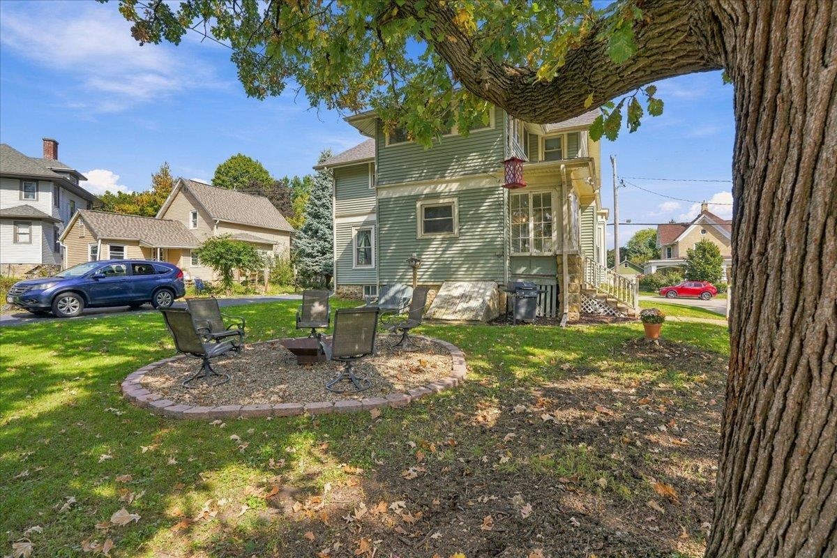 Rear of a green two-story house beside a shaded yard with a circular fire pit seating area.
