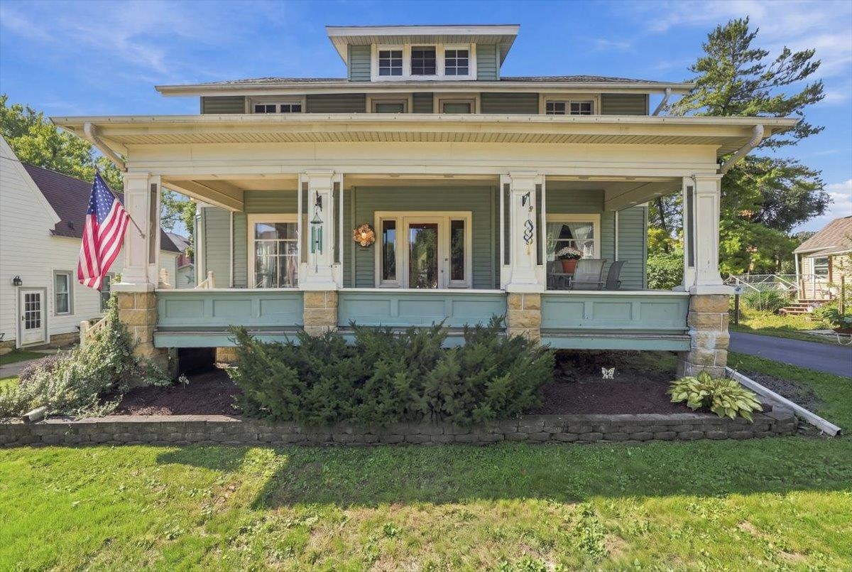 Green two-story house with a wide covered porch, stone supports, and an American flag.