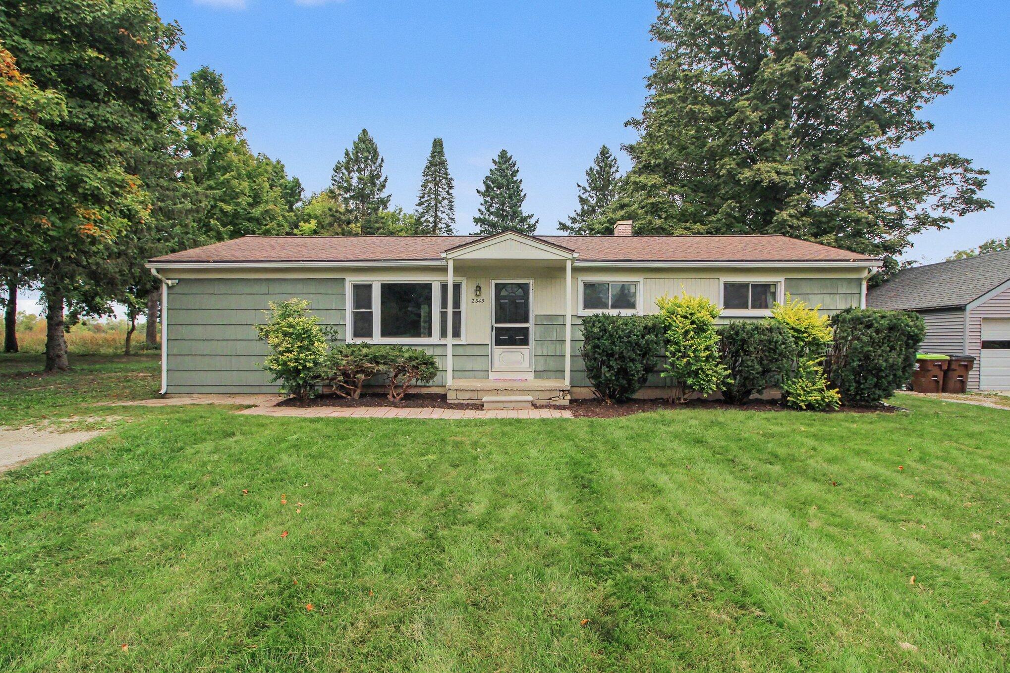 Single-story green ranch house with a covered entry, trimmed shrubs, and a broad front lawn.
