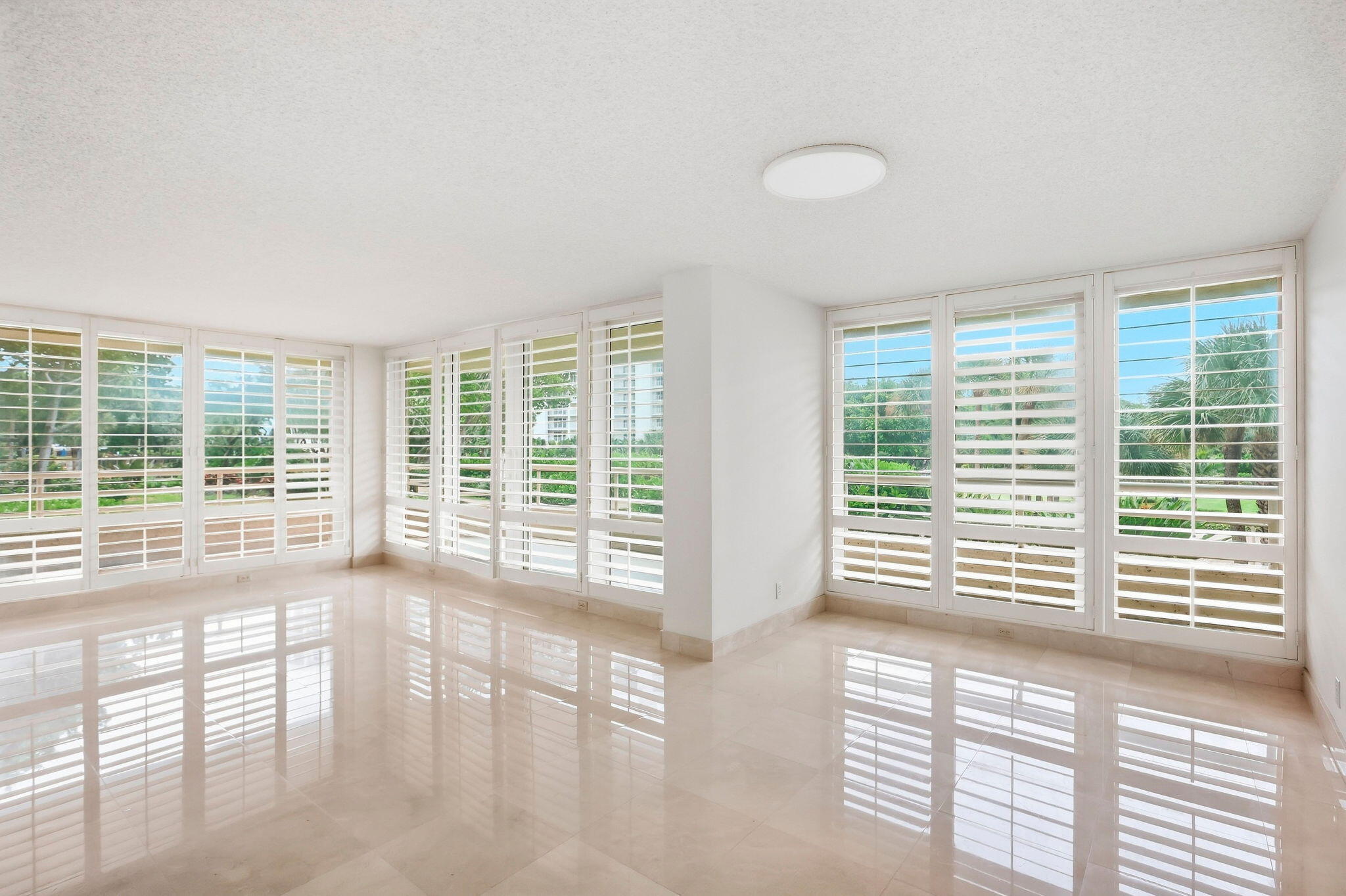 Vacant room with glossy tile floors and wraparound windows covered by white plantation shutters.