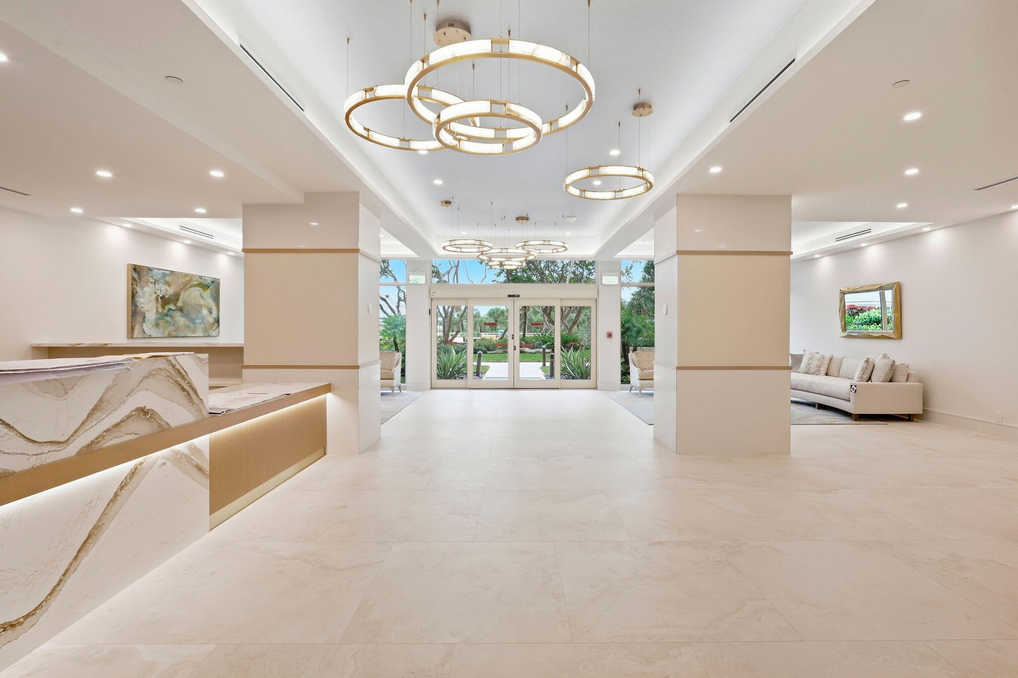 Condominium lobby with a marble reception desk, gold ring chandeliers, and glass doors overlooking greenery.