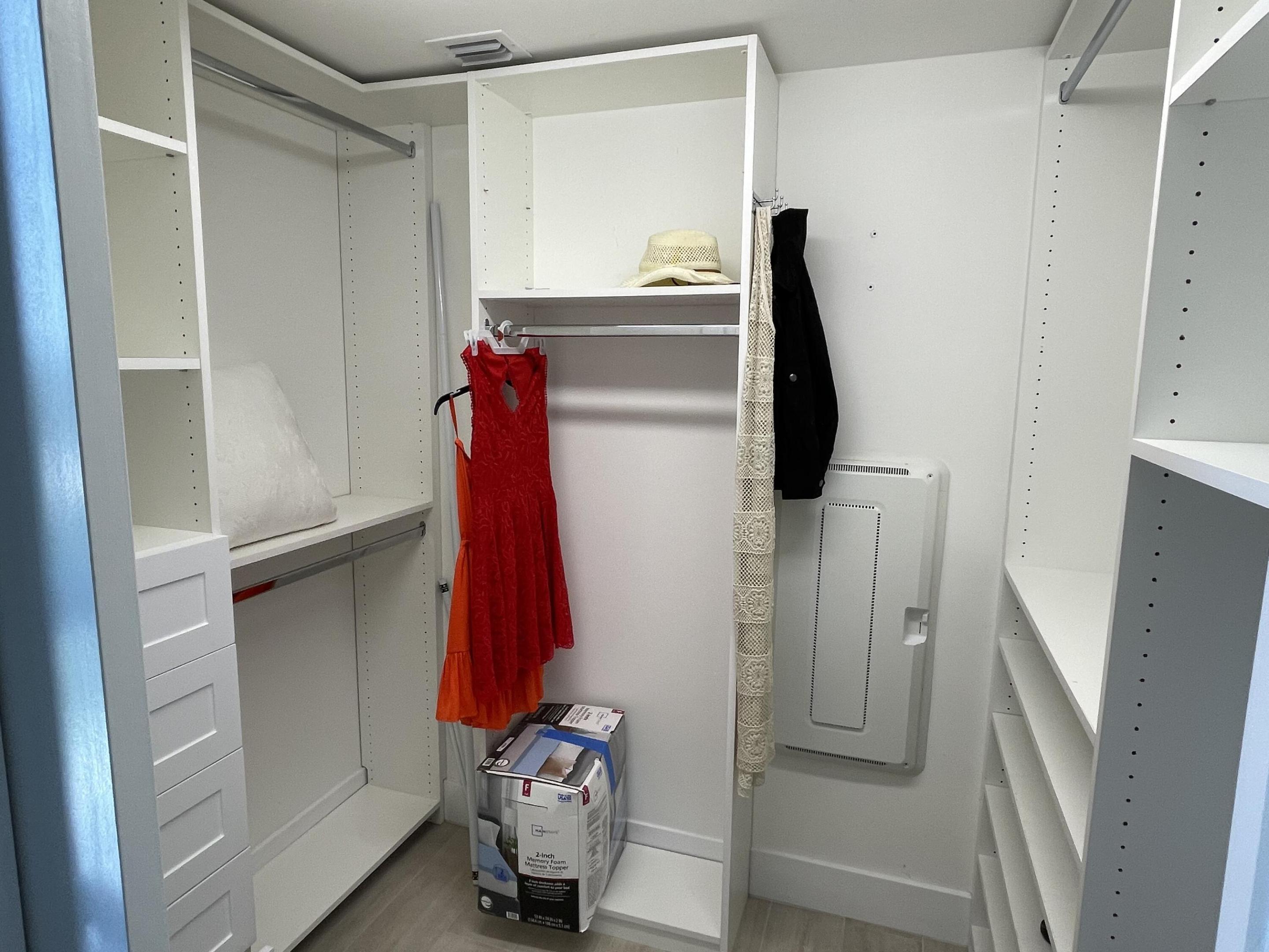 Walk-in closet with white built-in shelves and drawers, hanging clothes, and a mattress topper box on the floor.