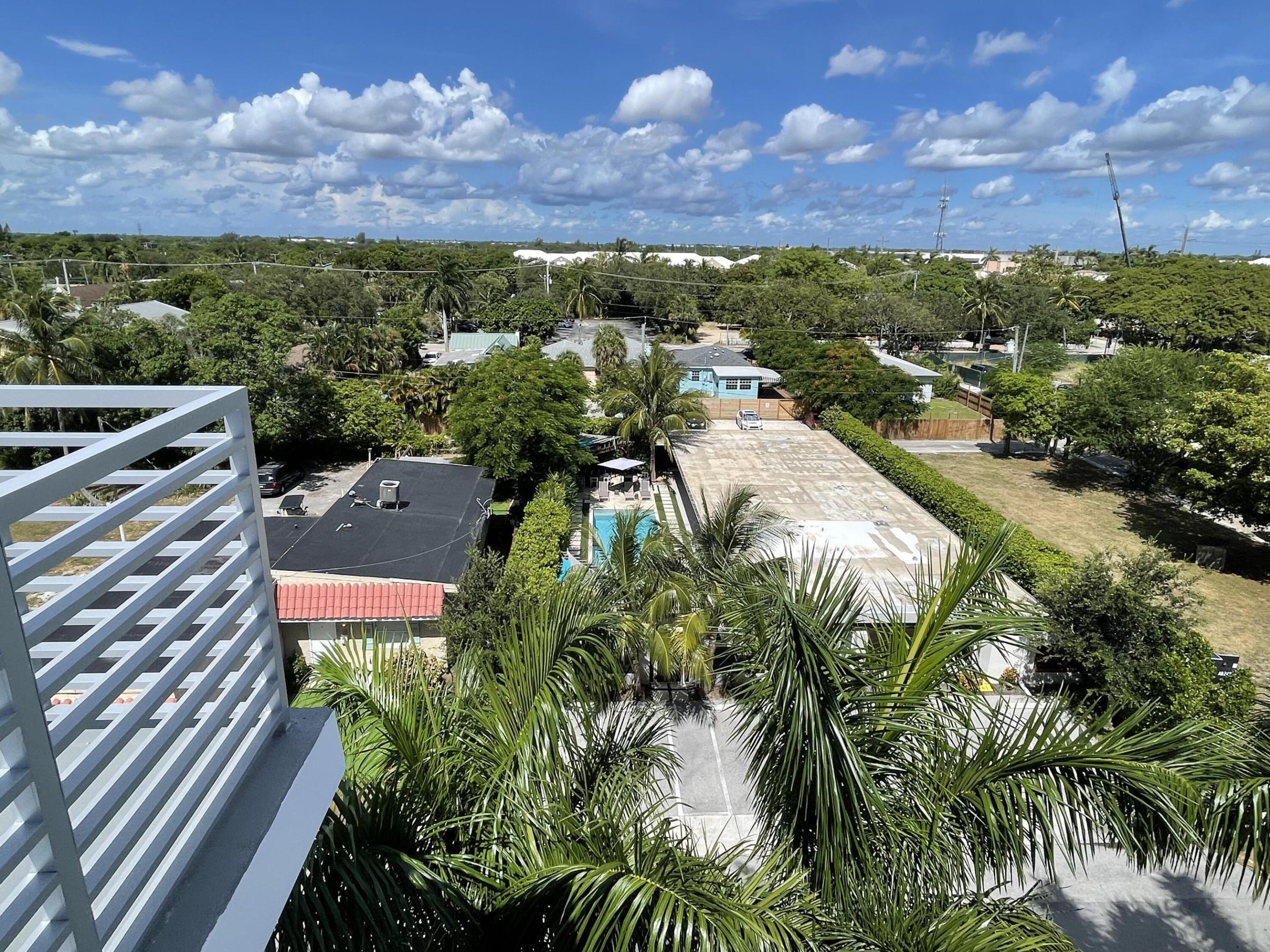 Elevated neighborhood view with palm trees, a swimming pool, and low-rise homes beneath a cloud-filled sky.