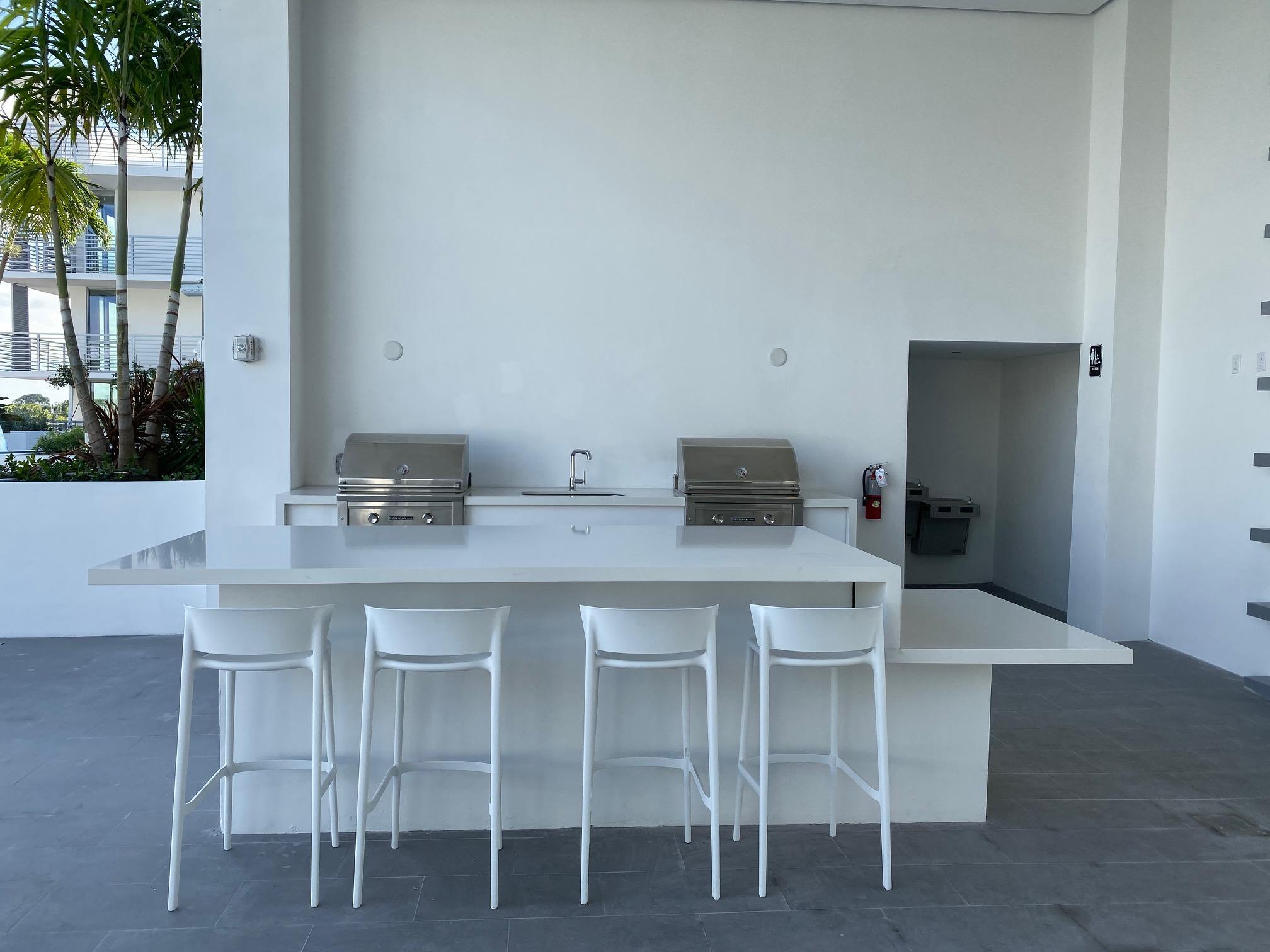 Covered outdoor kitchen with two grills, a sink, and a white bar island with four stools.